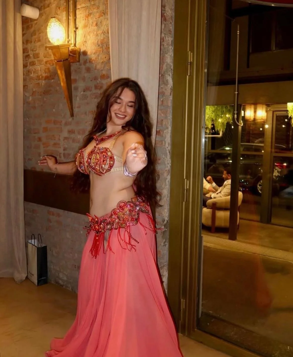 Una donna con capelli lunghi e ricci sorride e indossa un costume da belly dance con top decorato e gonna lunga rosa.