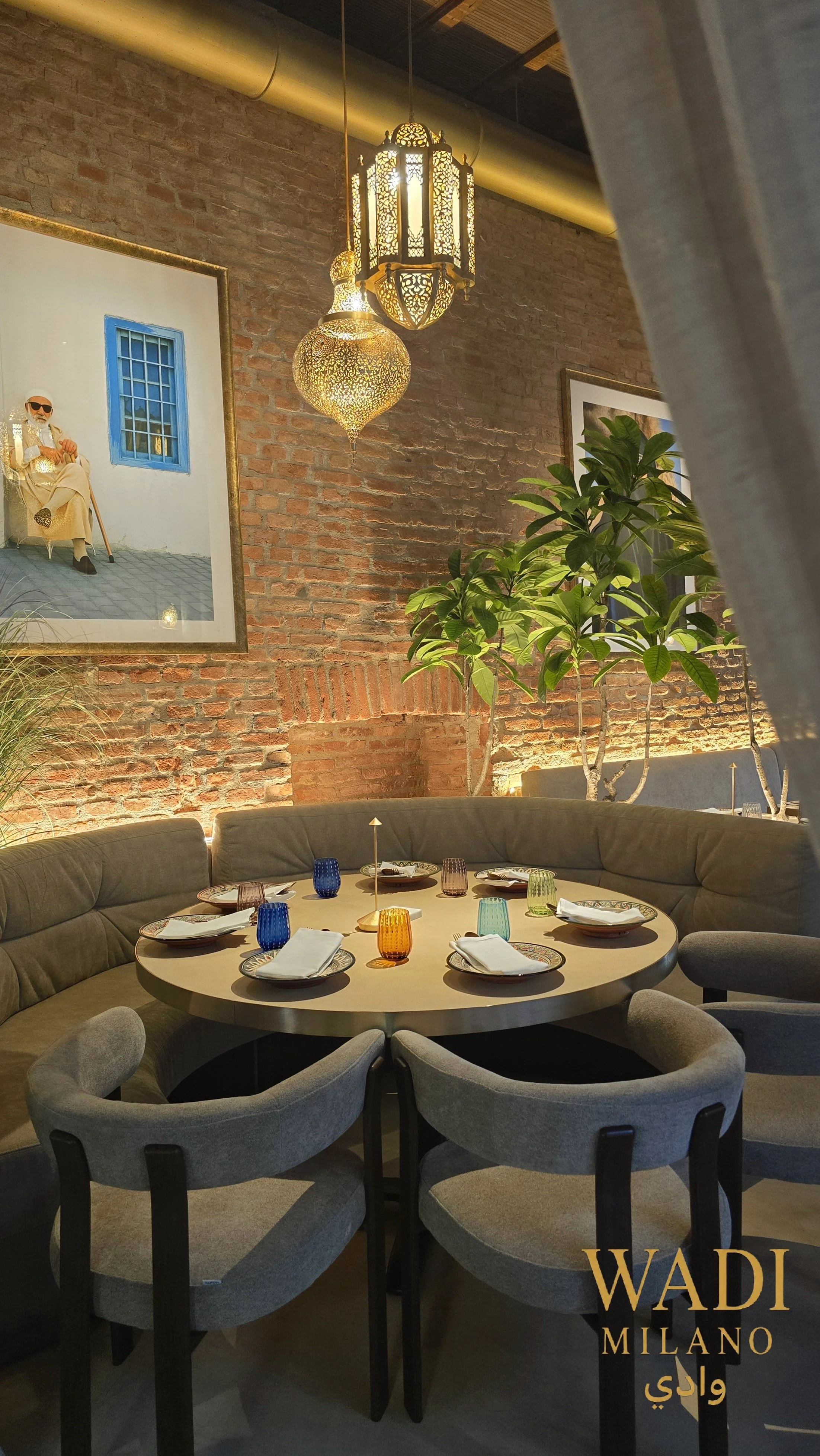 Interno di un ristorante elegante con pareti in mattoni a vista, tavolo rotondo apparecchiato con piatti, bicchieri colorati e posate, sedie imbottite grigie, piante verdi, lampade dai design etnici, e un quadro con figura umana e finestra blu, con i