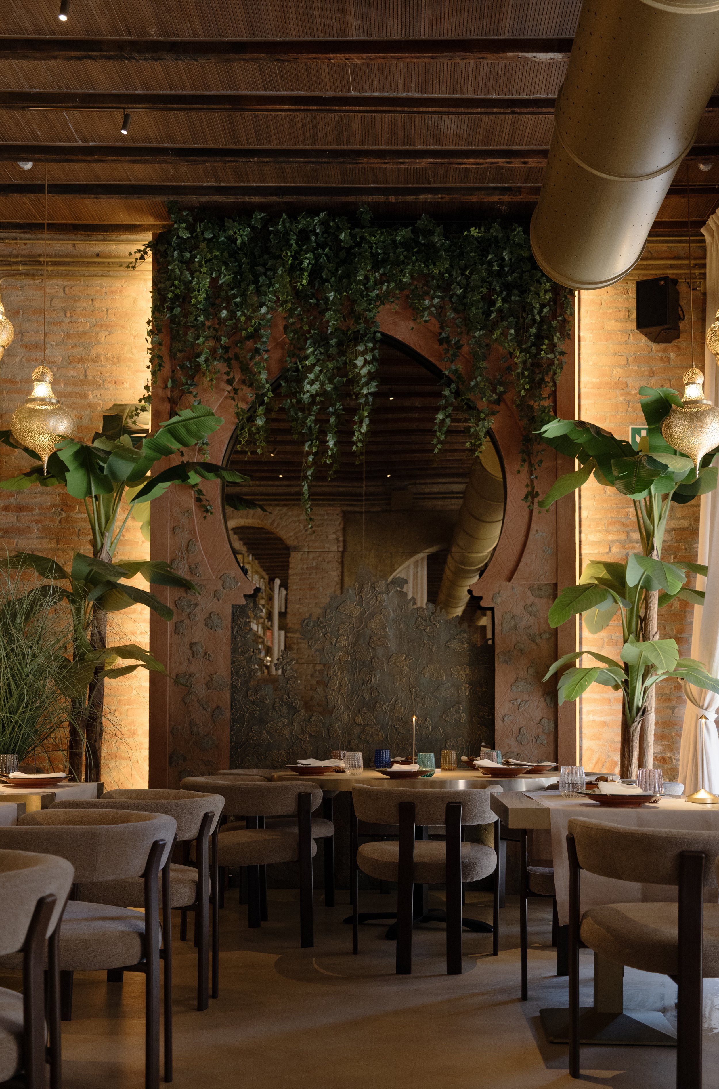 Interno di un ristorante elegante con pareti di mattoni, piante verdi e decorazioni luminose, tavoli apparecchiati con piatti, bicchieri e candele.