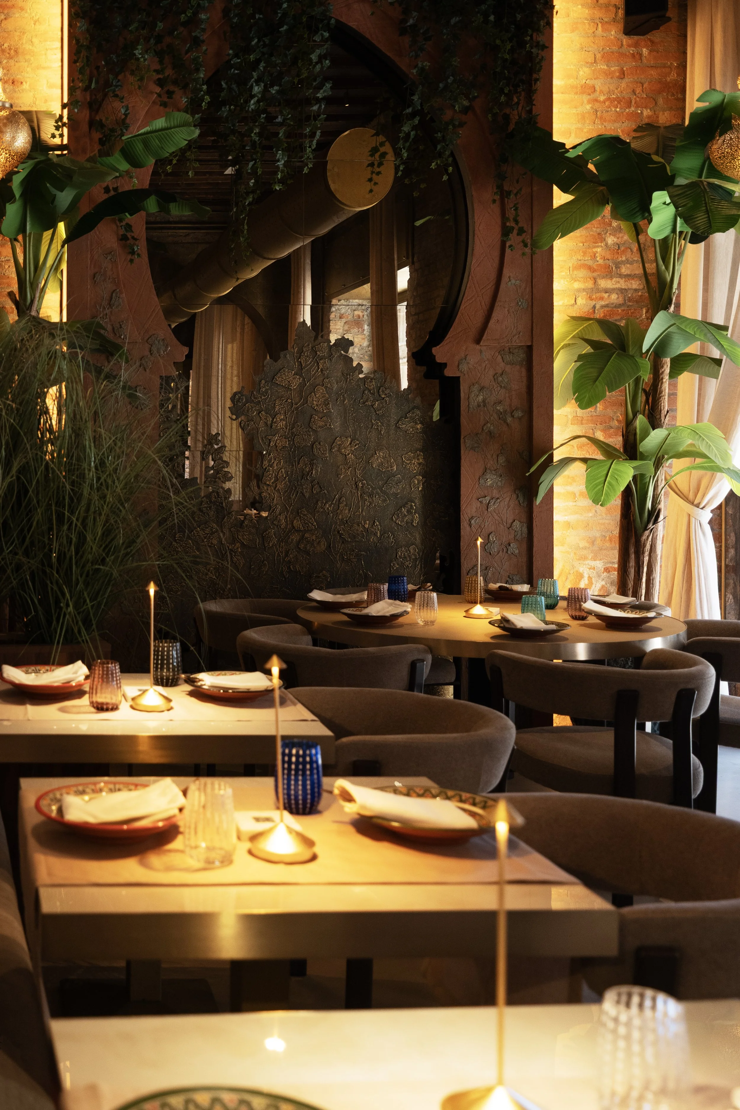 Interno di un ristorante elegante con tavoli apparecchiati, luci soffuse, piante verdi e pareti in mattoni.