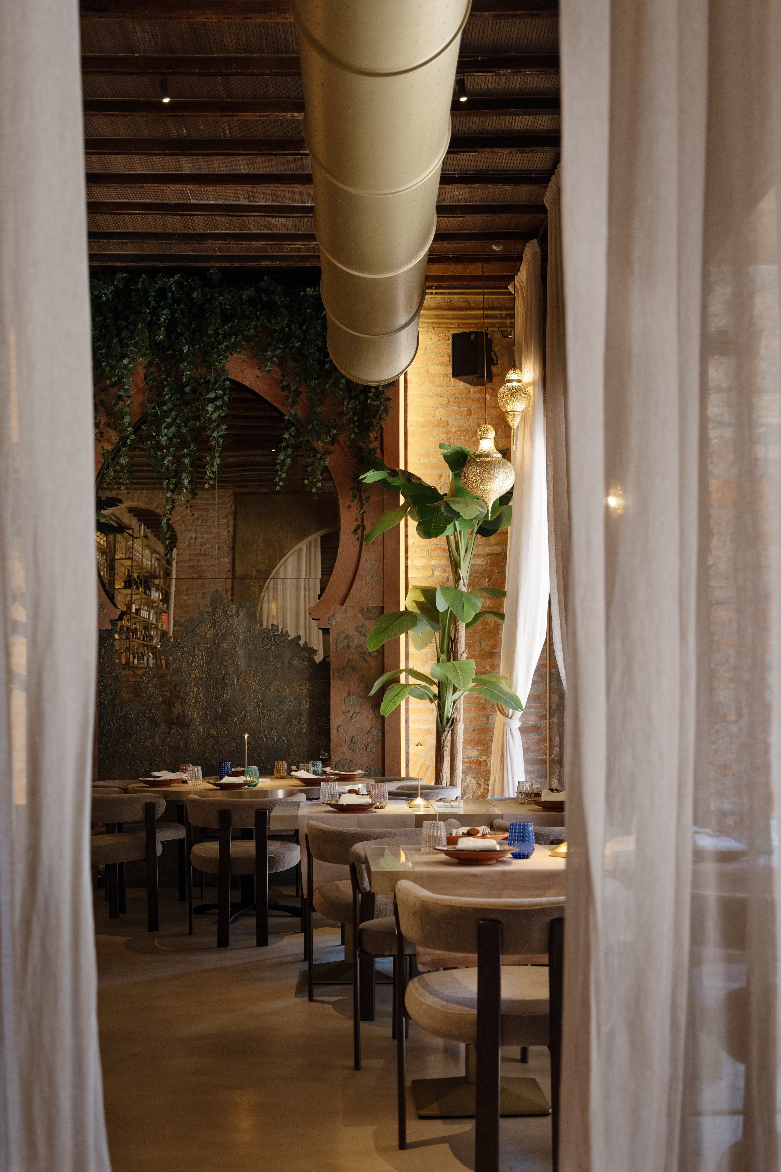 Interno di un ristorante elegante con tavoli apparecchiati, grandi piante e luci decorative, visti attraverso tende di tessuto leggero.