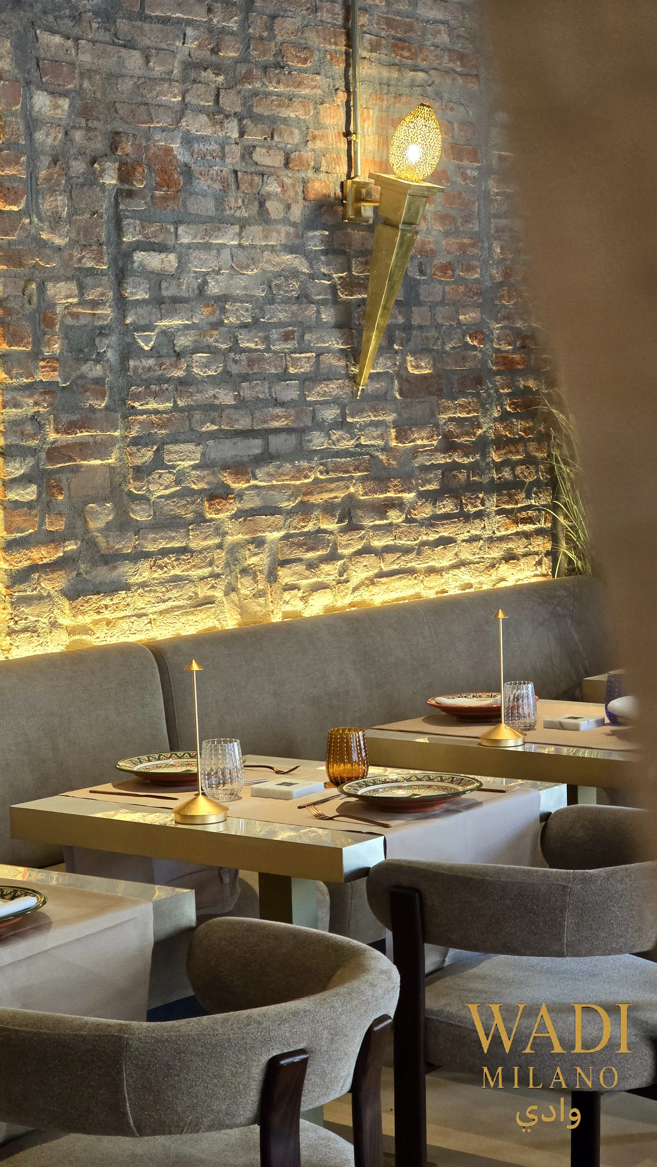 Interno di un ristorante elegante con tavoli apparecchiati, luci calde, un muro di mattoni a vista e decorazioni moderne, tra cui un'applique dorata e bicchieri e piatti raffinati sui tavoli.