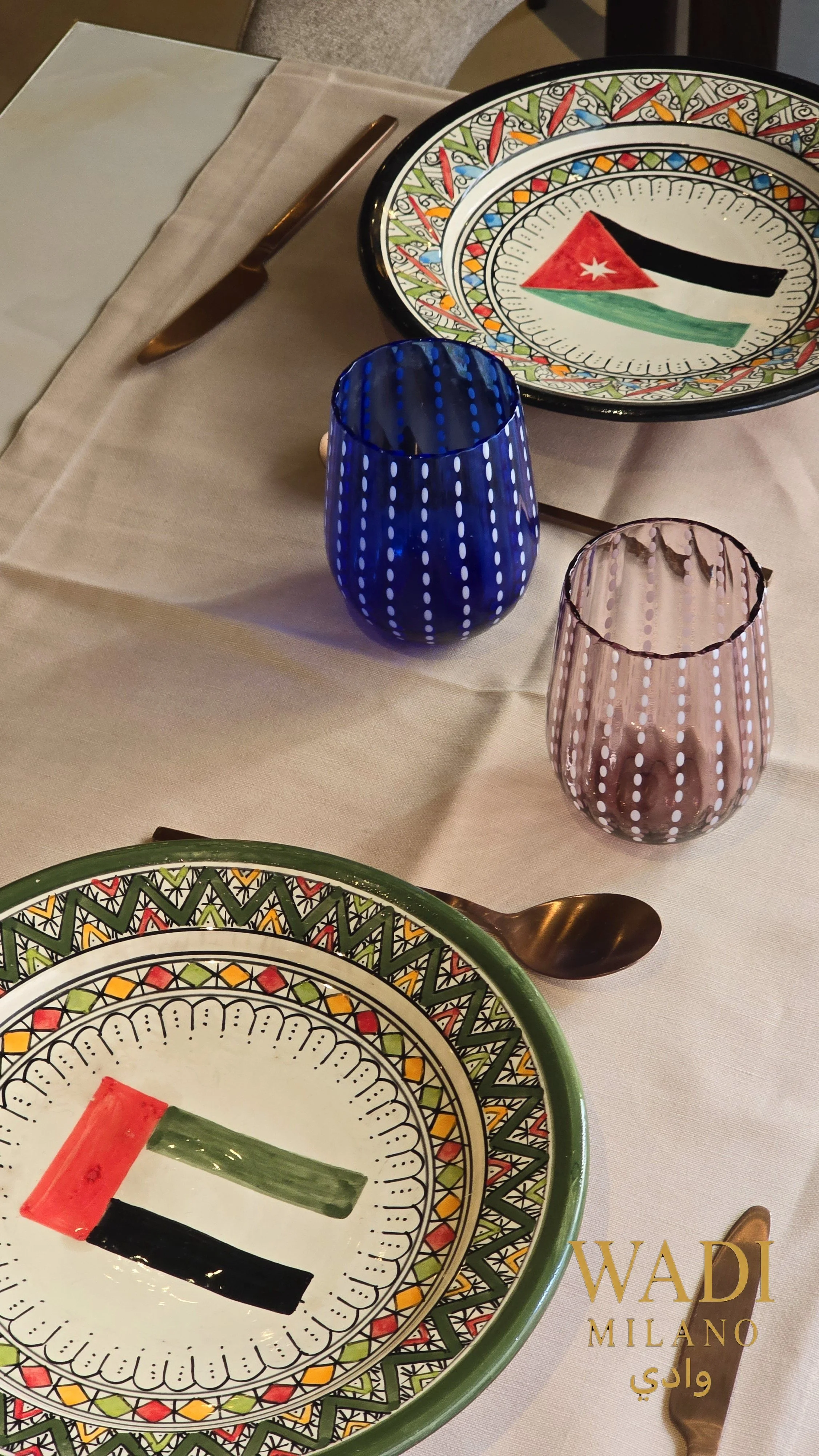 Piatti colorati con design arabo, bicchieri blu e rosa, posate d'argento, tovaglia beige, e logo Wadi Milano scritto in oro.
