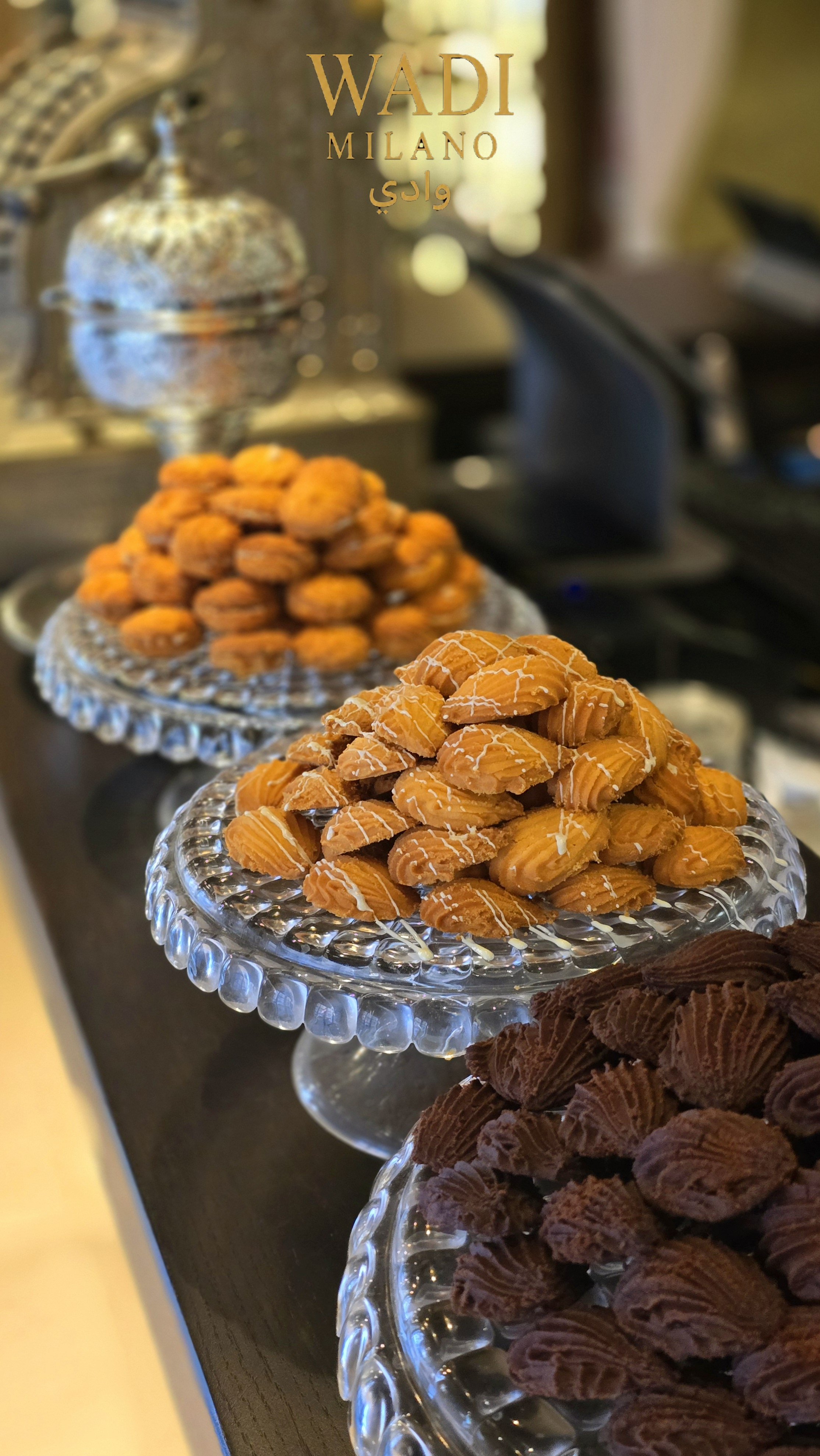 Tre vassoi con vari tipi di dolciumi su un tavolo, in primo piano ci sono biscotti con glassa bianca, poi ci sono altri dolci arancioni e cioccolato fondente.
