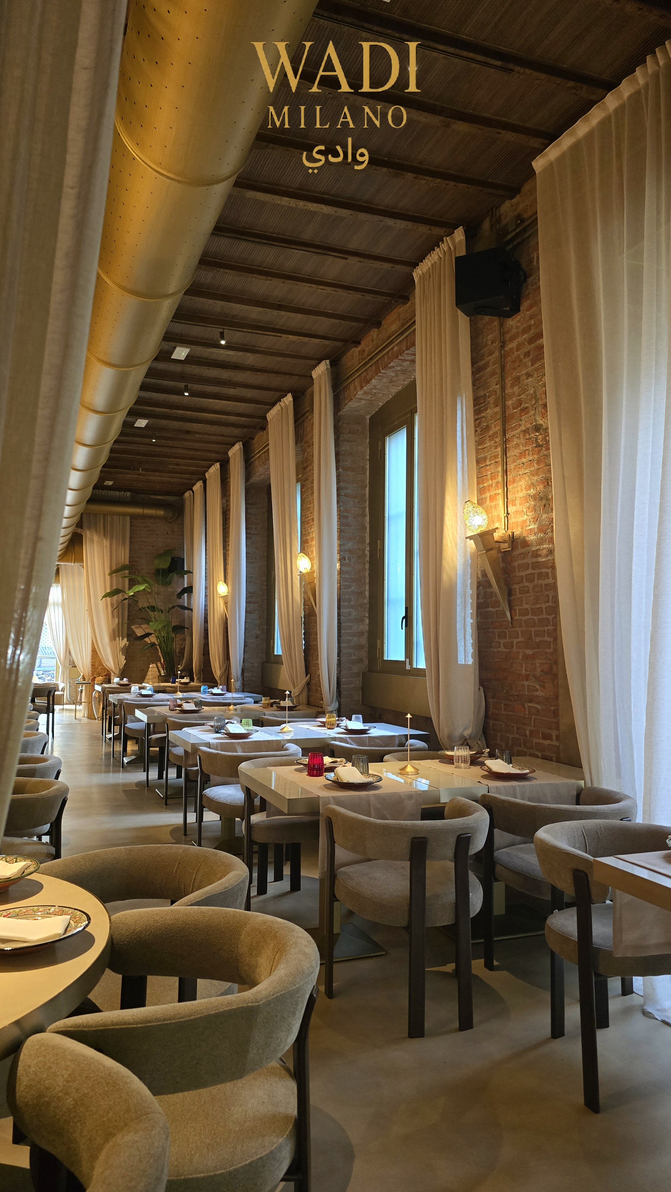 Interno di un ristorante elegante con tavoli apparecchiati, grandi finestre con tende bianche e pareti di mattoni rossi, illuminazione calda e decori moderni.