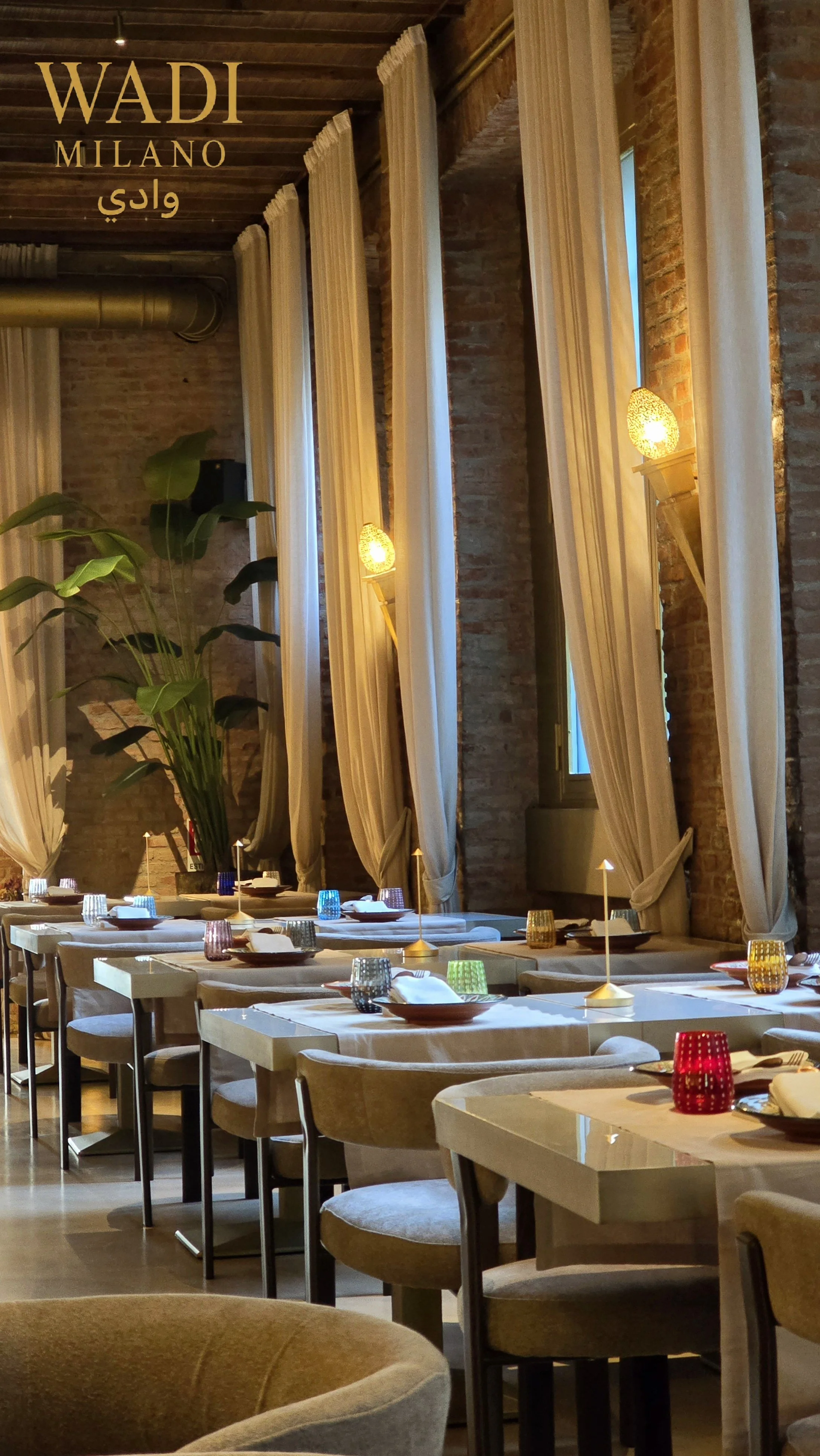 Interno di un ristorante elegante con tavoli aperti, tovaglie bianche, bicchieri colorati, candele e grandi tende beige, pareti in mattoni e luci calde.