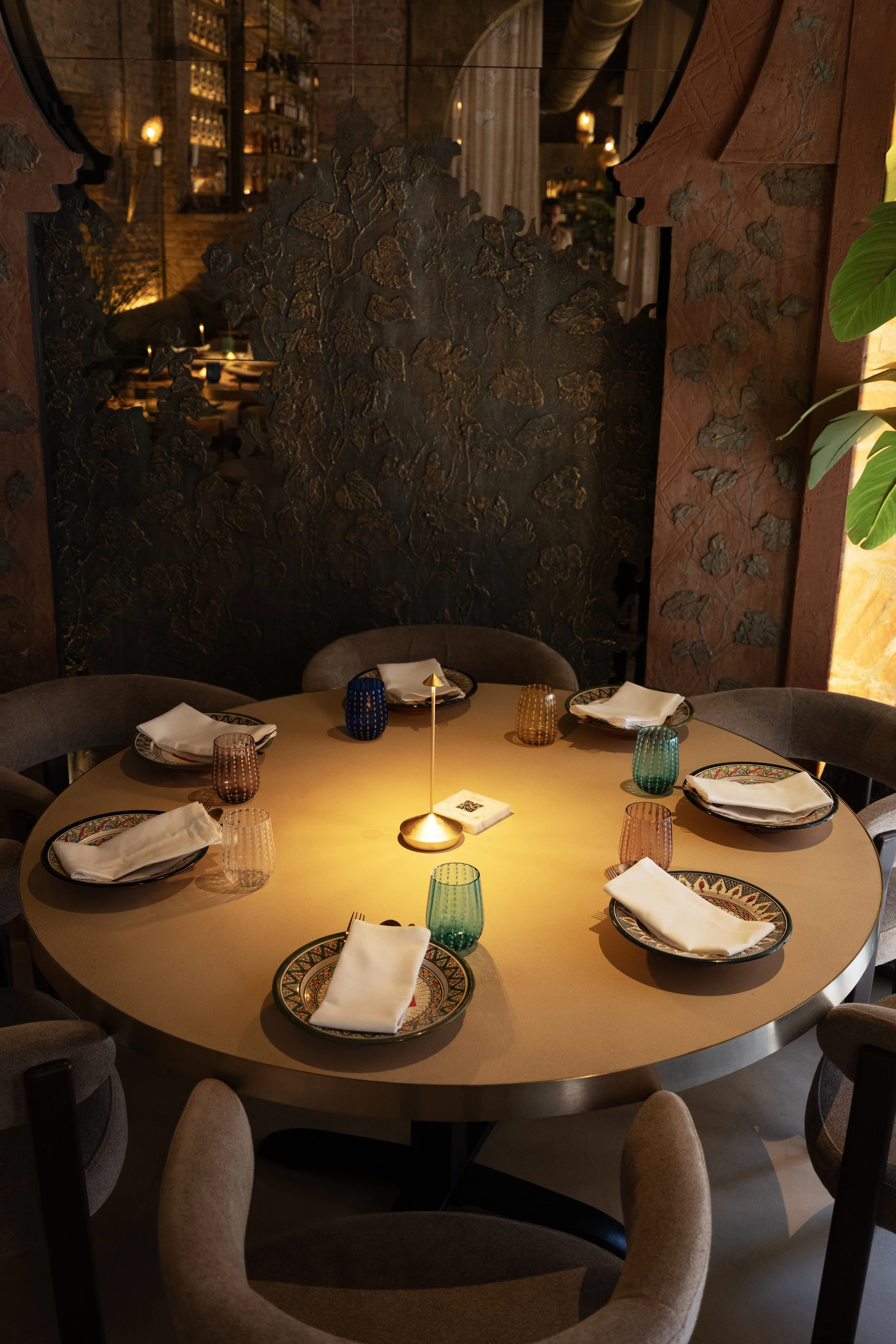 Una tavola apparecchiata con piatti, bicchieri colorati e tovaglioli bianchi in un ristorante con decor rustico, pareti in mattoni e giochi di luce calda.
