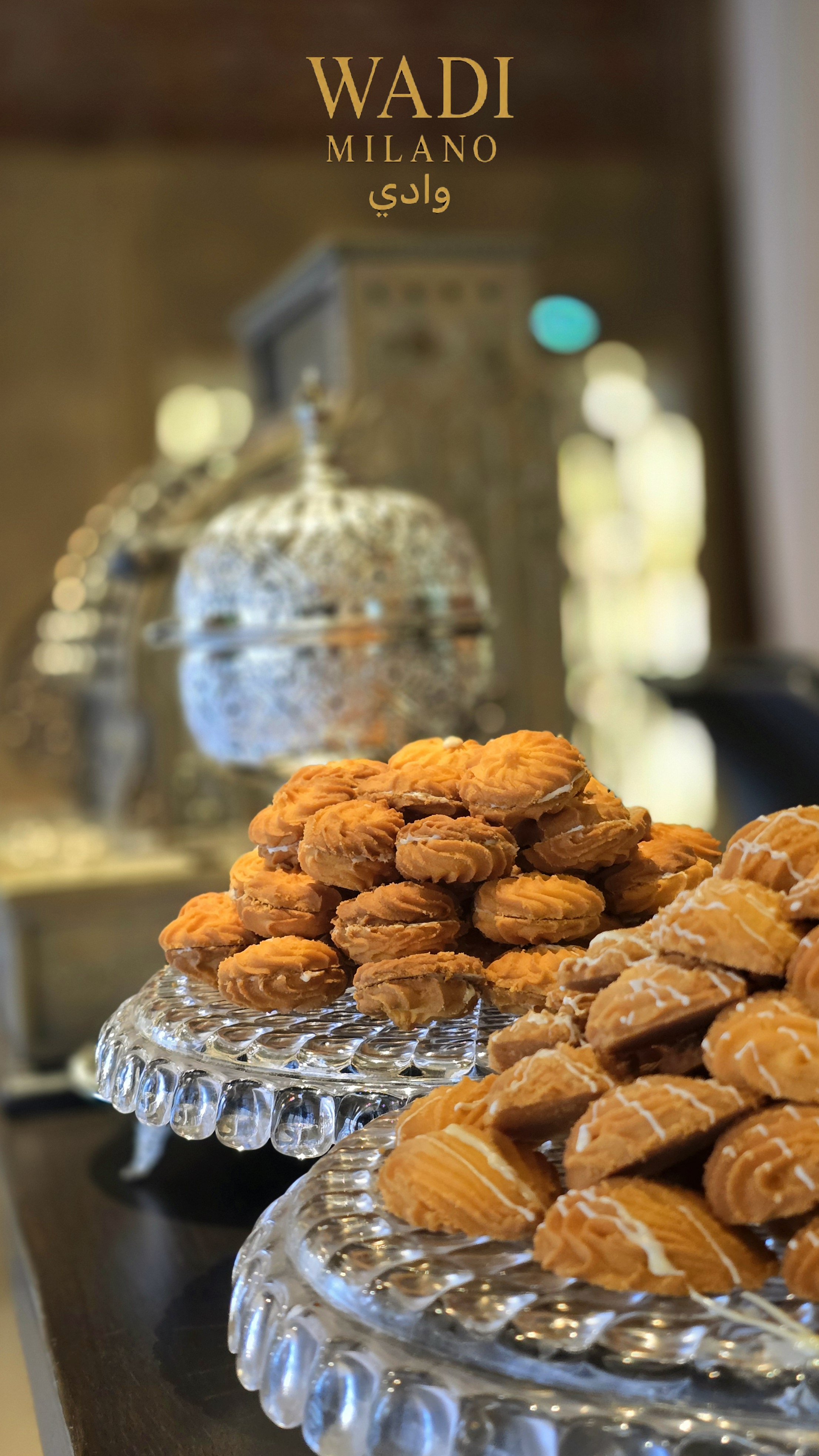 Desserti e biscotti posizionati su piatti di vetro su un tavolo, con sfondo sfocato. In evidenza ci sono biscotti di vari tipi, alcuni decorati con glutine o glassa.