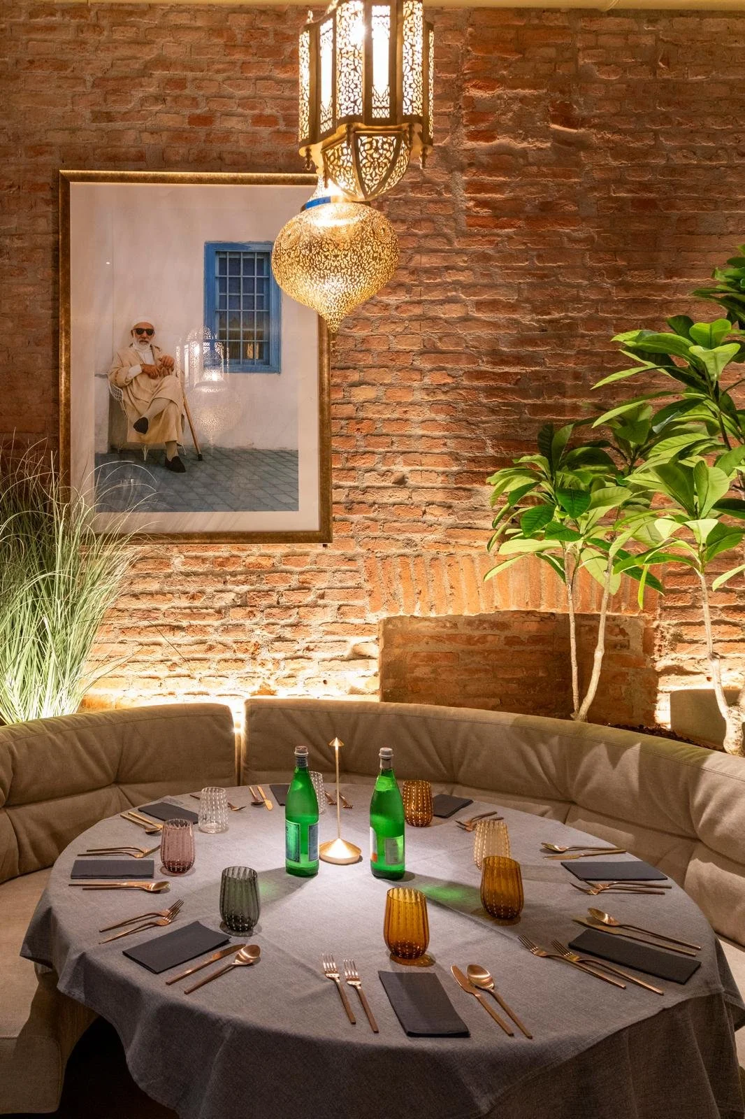 Una sala da pranzo elegantemente arredata con un tavolo rotondo coperto da una tovaglia grigia, apparecchiatura in oro rosa, bicchieri colorati e due bottiglie di acqua. La stanza ha pareti di mattoni rossi, piante verdi e un ritratto di uno uomo con barba, occhiali e vestiti chiari appeso alla parete. L'illuminazione calda crea un'atmosfera accogliente.
