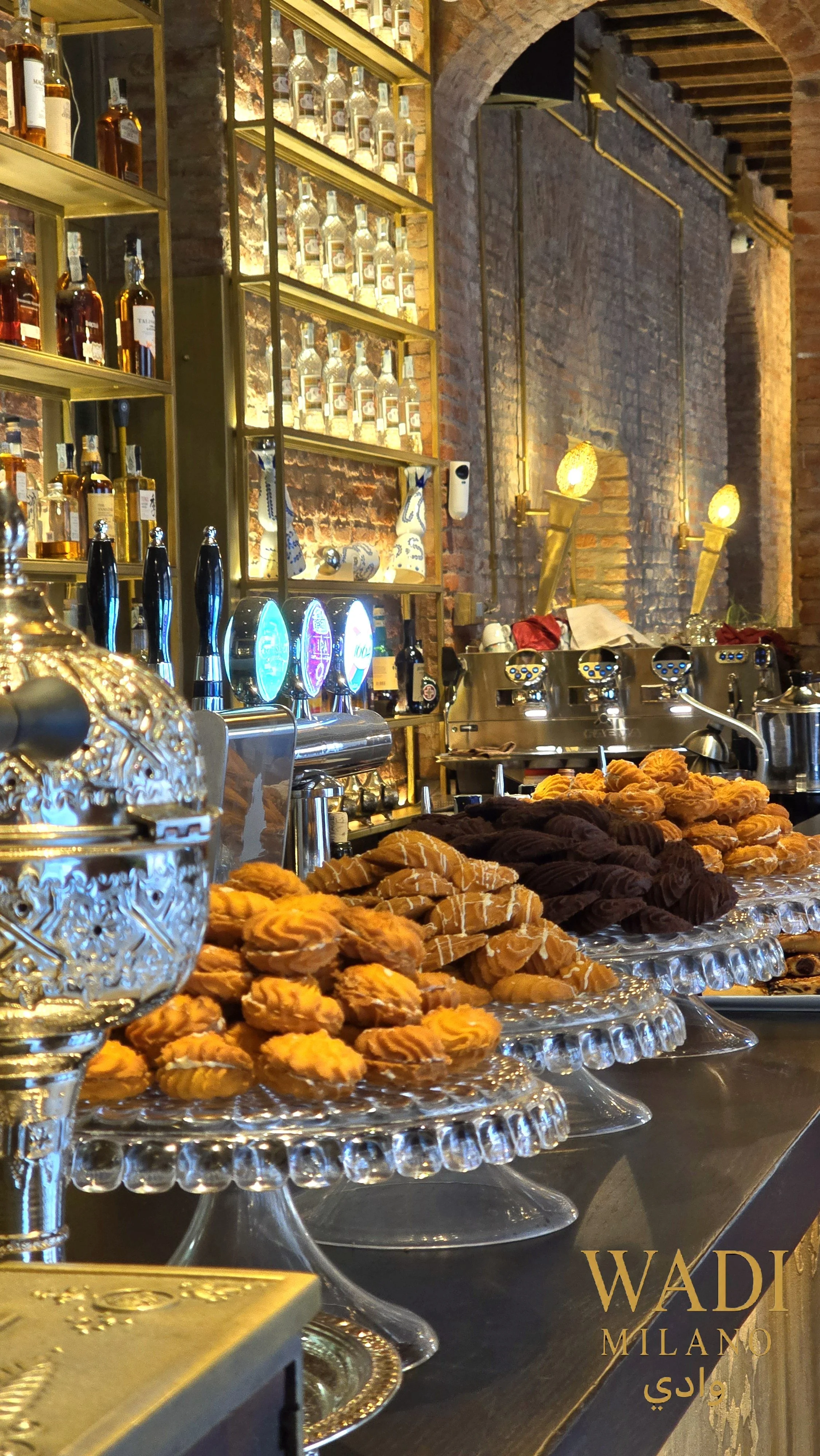 Una scena di bar con scaffali pieni di bottiglie di alcolici, luci calde e decorazioni eleganti, con dolciumi su vassoi di vetro in primo piano.