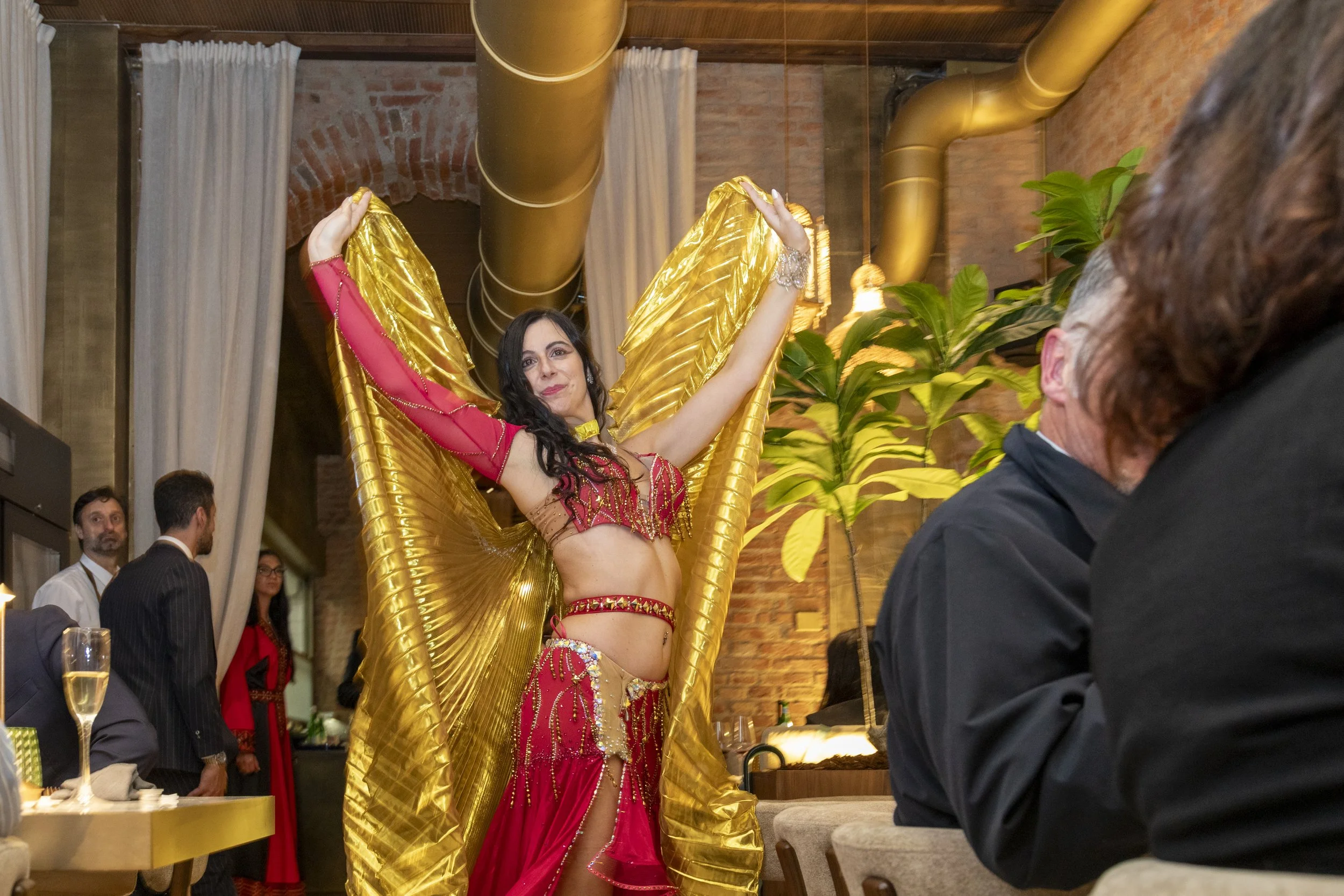 Una donna vestita con un costume da spettacolo rosso e oro, con grandi ali dorate, che danza durante un evento in un ristorante o sala da feste, con persone sedute e che osservano, piante e elementi decorativi in legno e mattoni sullo sfondo.