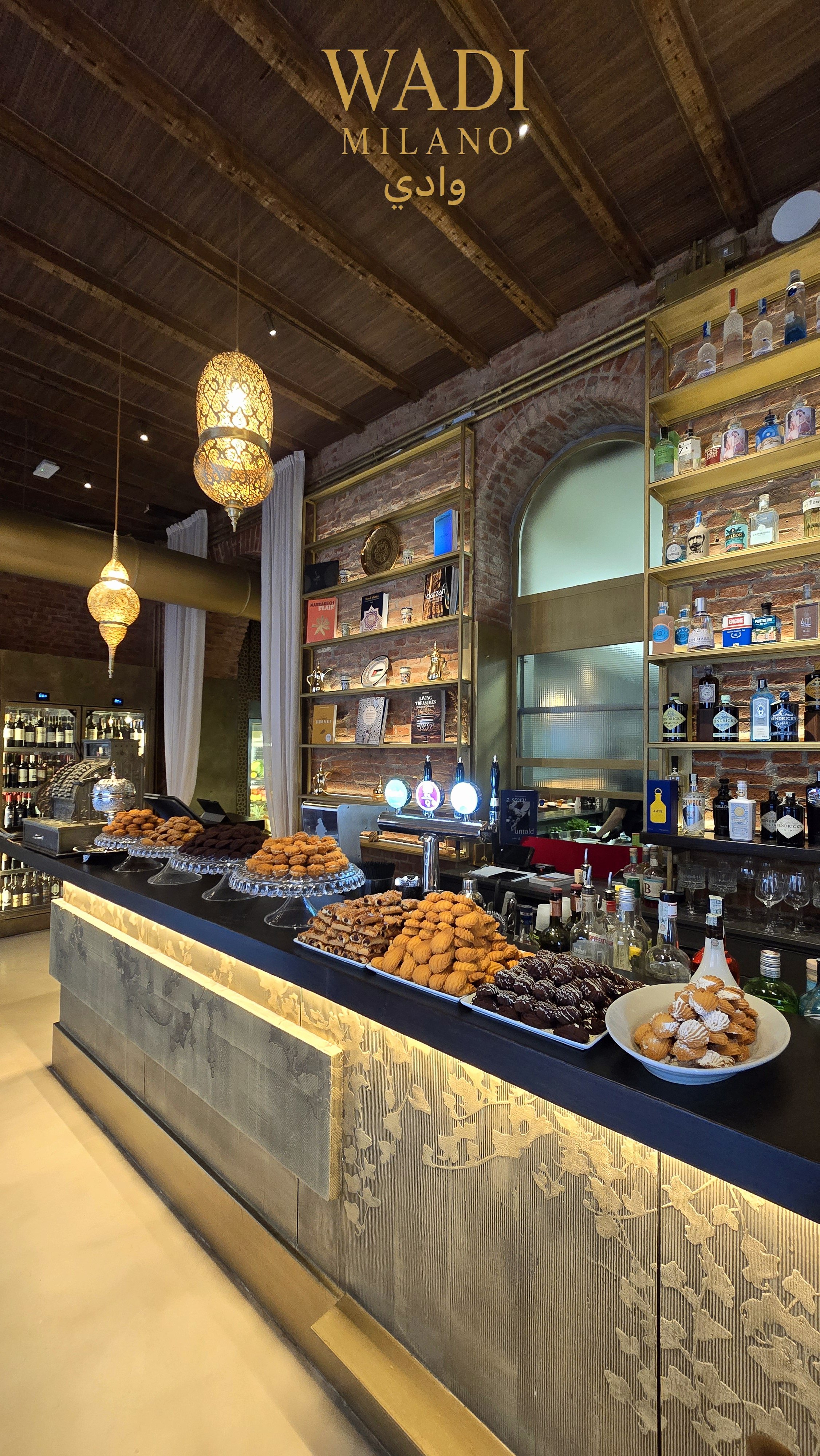 Un bar con scaffali di bottiglie di alcolici, un bancone con vari dolci e snack, lampade decorative, muri in mattoni e un'area con tende bianche.