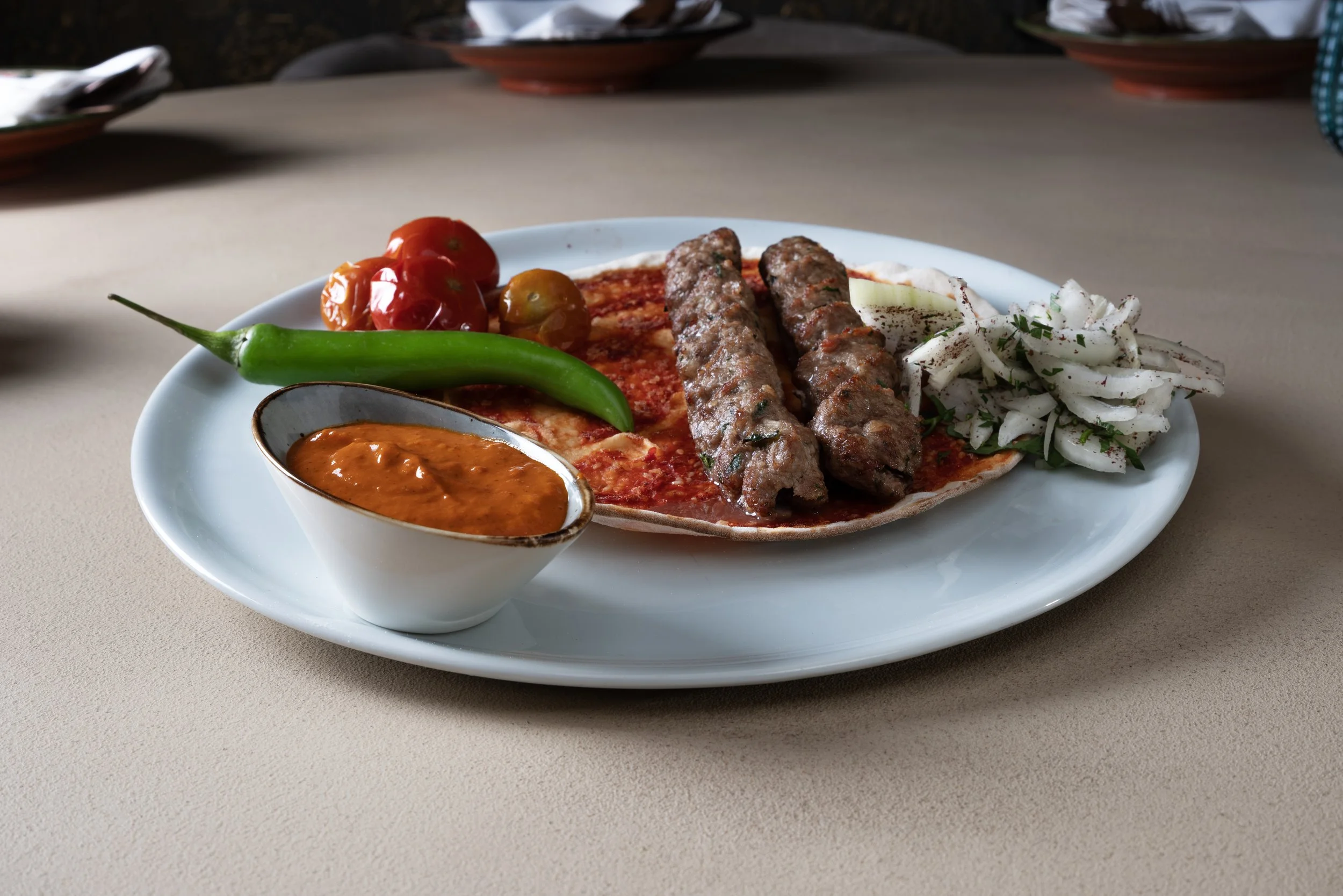 Piatti di cibo con due spiedini di carne, insalata di cipolla, pomodori ciliegia, un peperoncino verde e una ciotola di salsa piccante, su piatto bianco su tavolo beige.