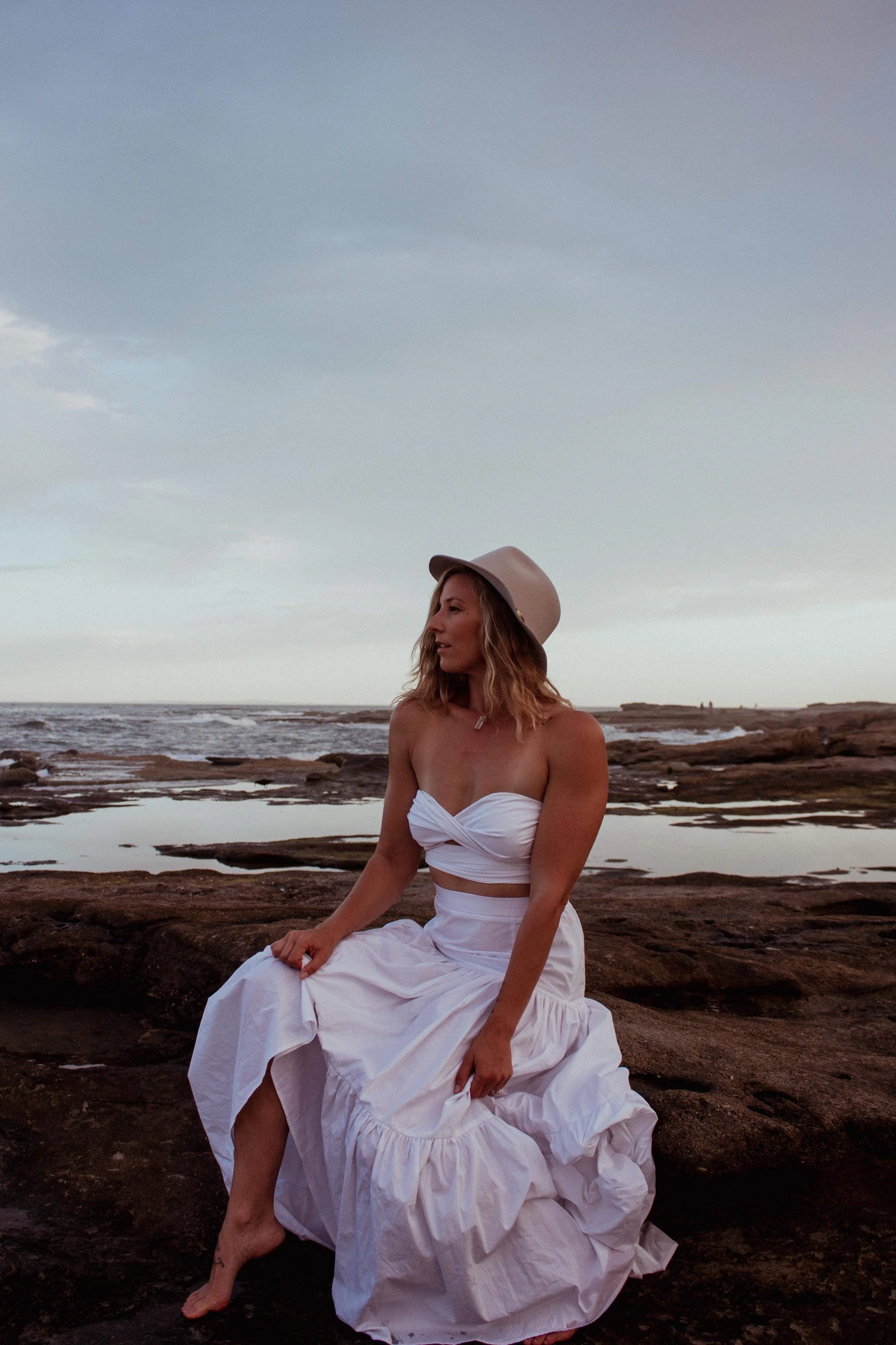 A woman sitting on rocks at the beach, wearing a white strapless dress and a beige hat, with an overcast sky and the ocean in the background.