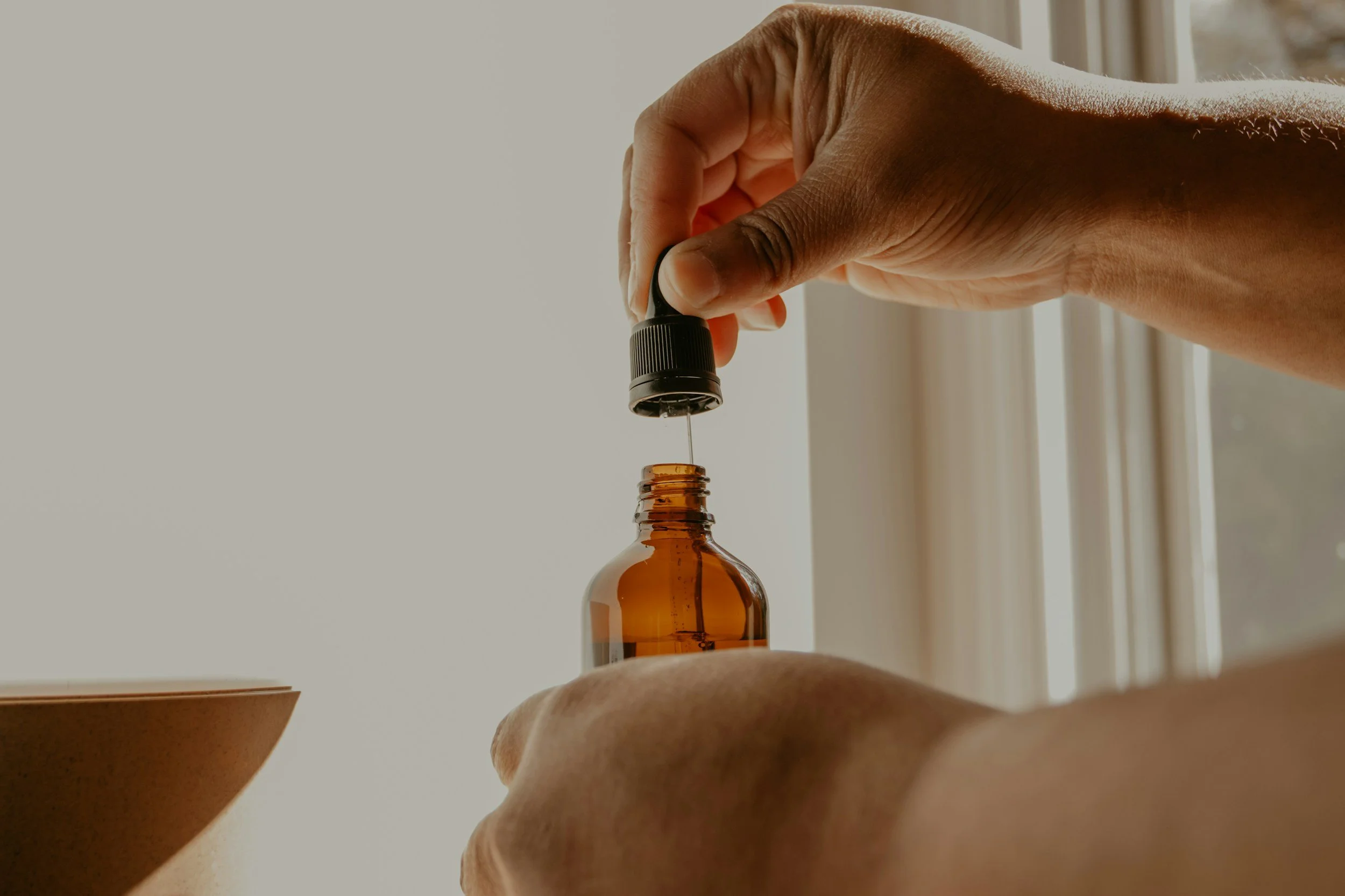 A person is dropping essential oil from a dropper into a glass bottle on a wooden surface, with another bowl nearby.