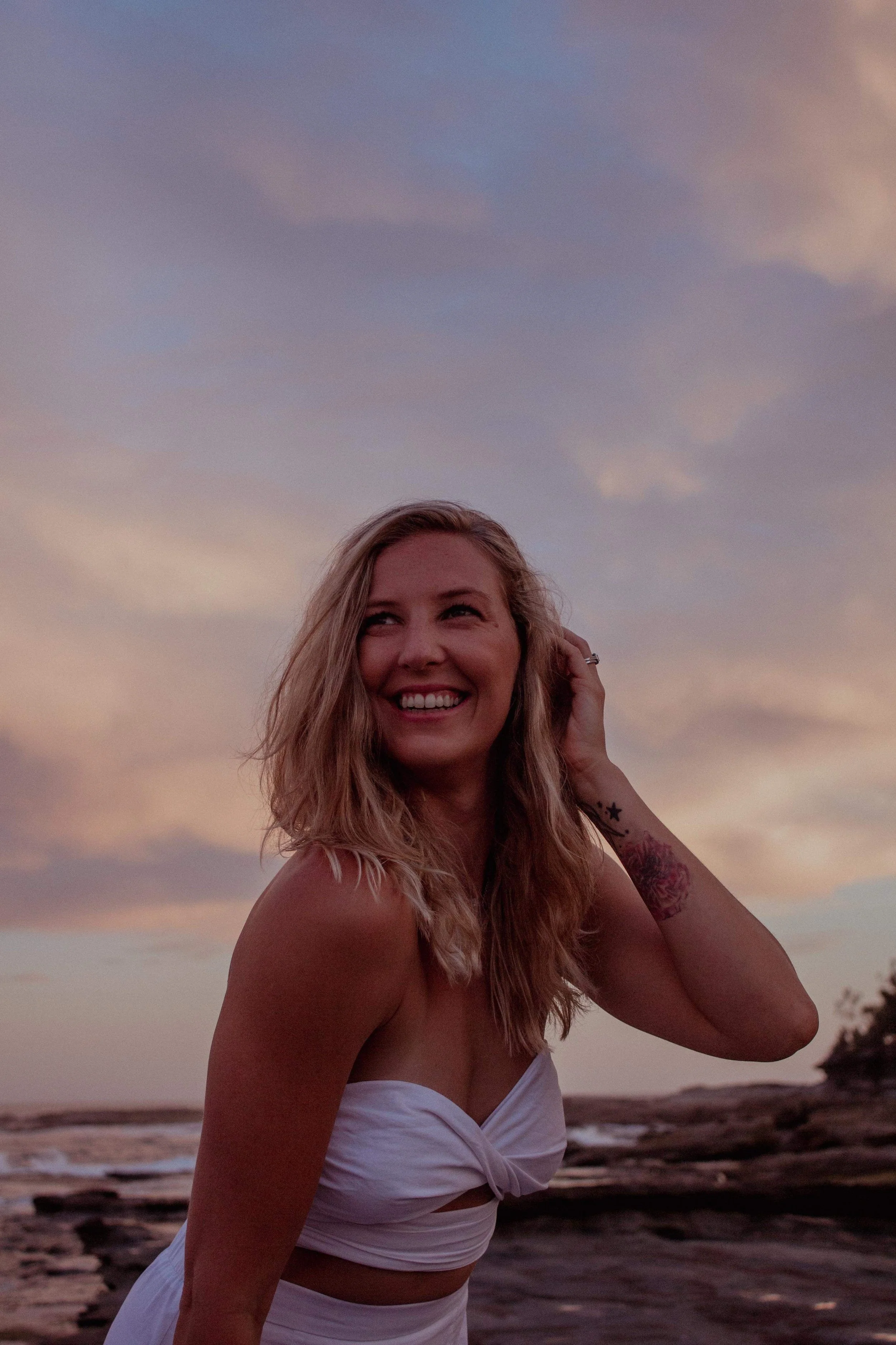 A woman smiling with long blonde hair, wearing a white strapless dress, standing near a rocky shoreline at sunset or sunrise with a cloudy sky overhead.