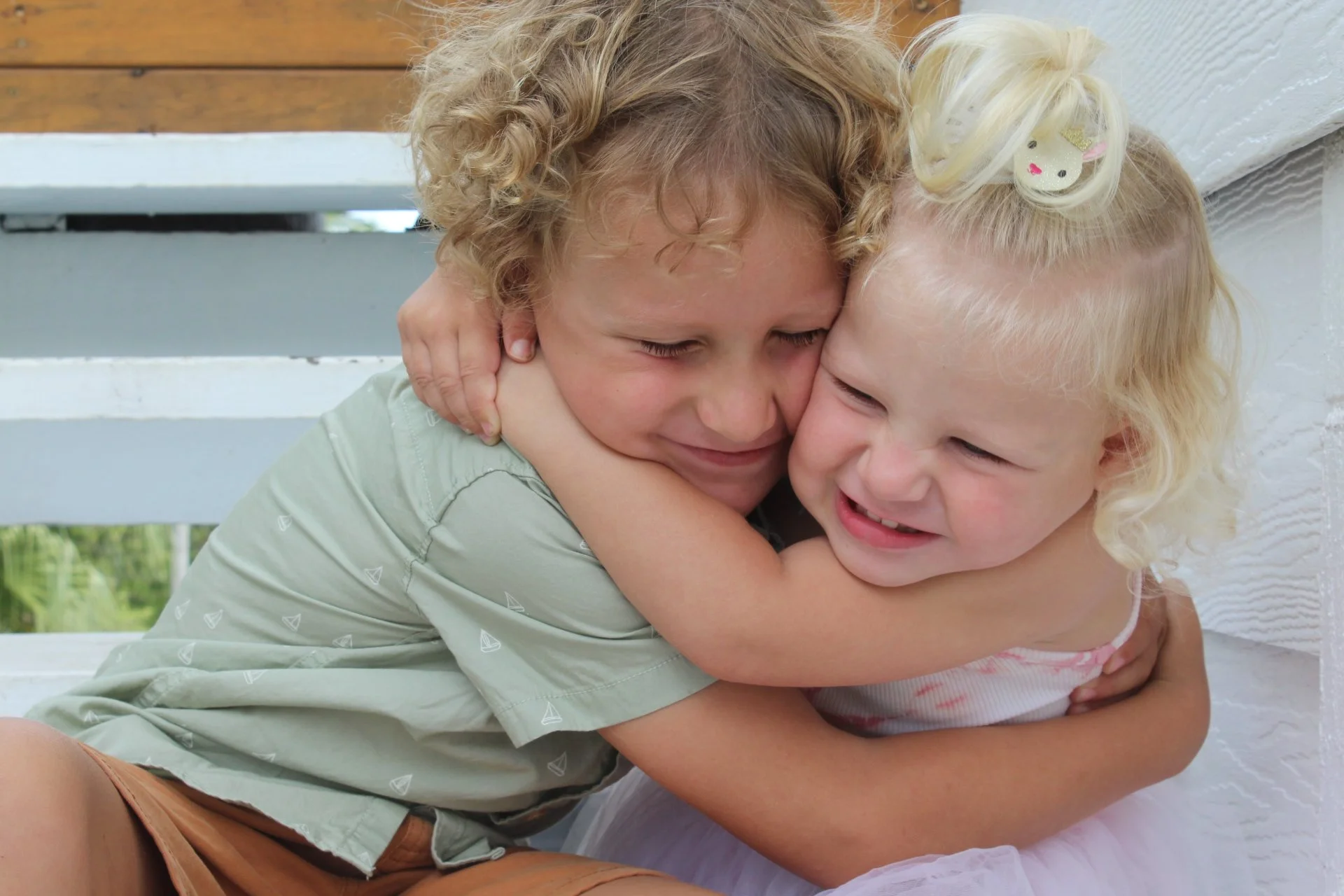 Two young children hugging each other tightly with happy expressions. One has curly blonde hair and a decorative hair clip, the other has straight blonde hair. They are outside, near a white wooden bench.