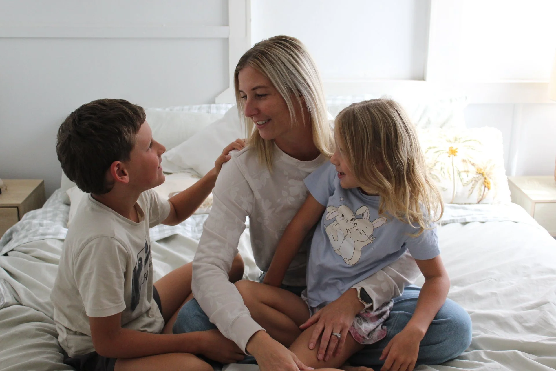 A woman and three children sitting on a bed, smiling and engaging in a conversation in a bright bedroom.