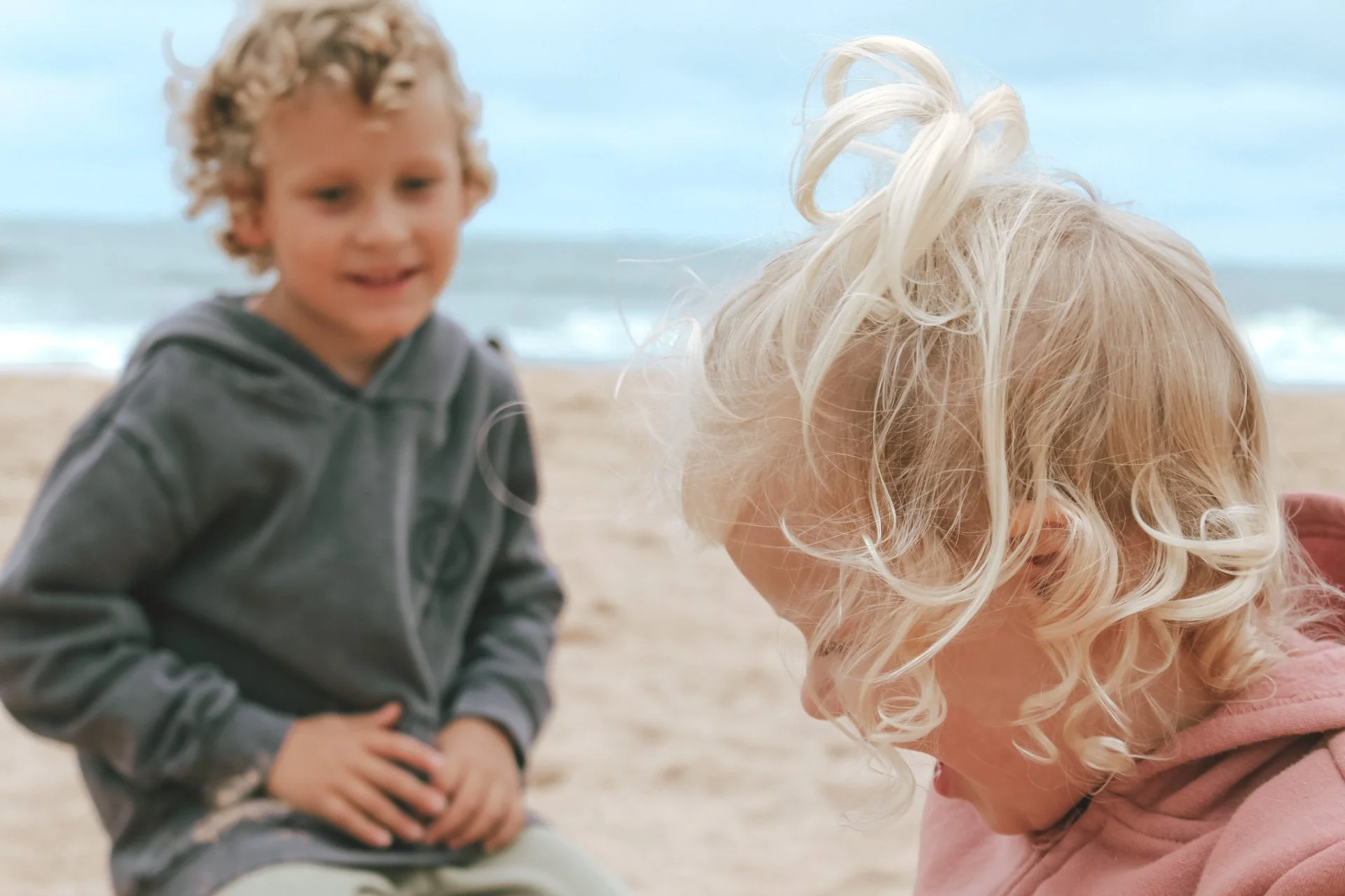 Two children on a beach, one with curly blond hair leaning forward, and another with other children blurred in the background, standing and wearing a dark hoodie.