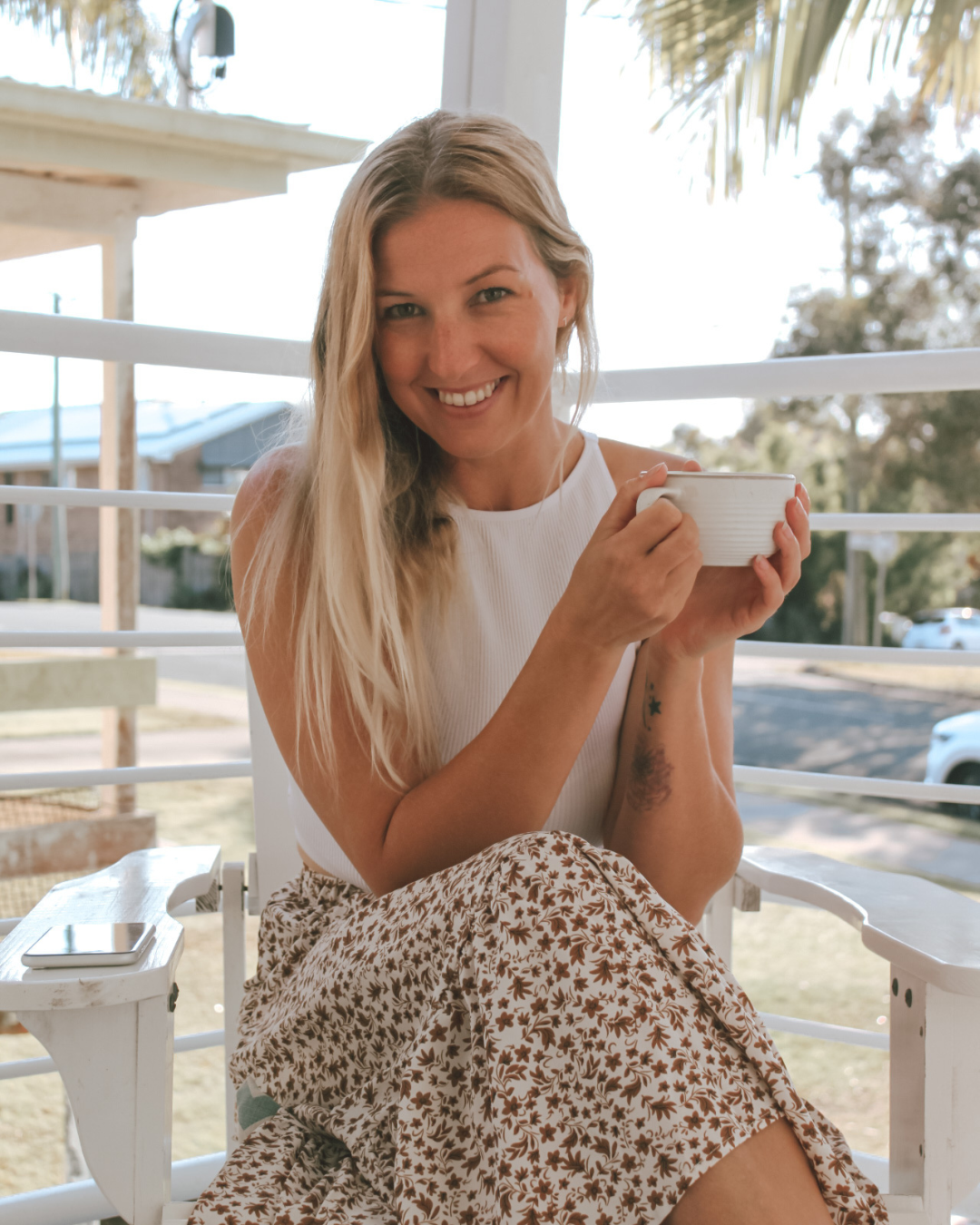 A smiling woman with long blonde hair sitting on a white porch railing, holding a white mug, wearing a sleeveless white top and a patterned skirt, with a smartphone placed on the armrest beside her, outdoors during daytime.