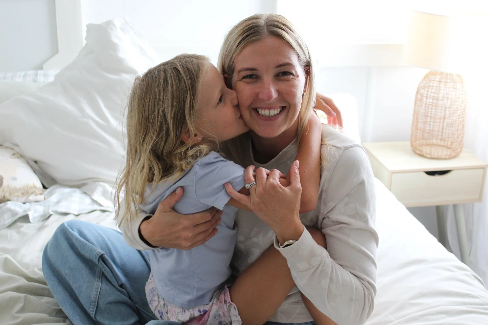 A woman and a young girl, possibly mother and daughter, sitting on a bed in a bright bedroom, embracing and sharing a kiss, smiling happily.