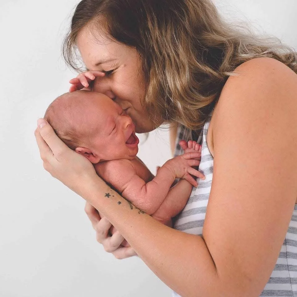 A woman with brown hair holding and kissing a crying newborn baby.