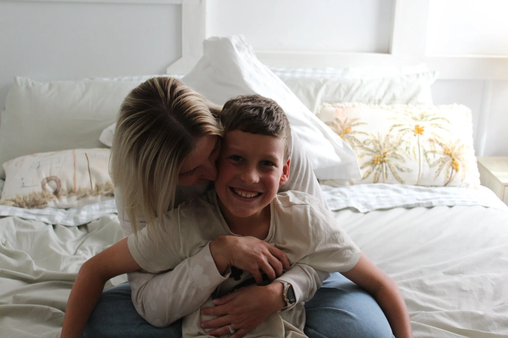A woman and a boy laughing and playing on a bed in a bright, white room.