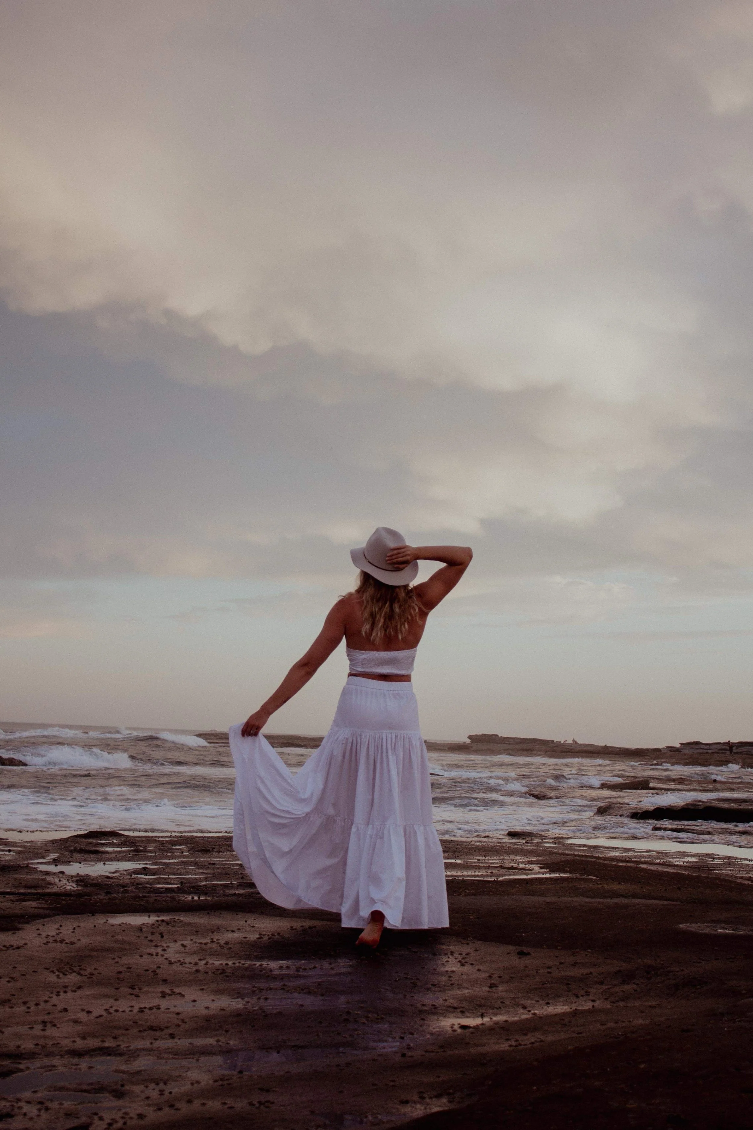 A woman in a white dress and a wide-brimmed hat standing on a beach at sunset, with her back to the camera, holding her dress with one hand and gazing out at the ocean.