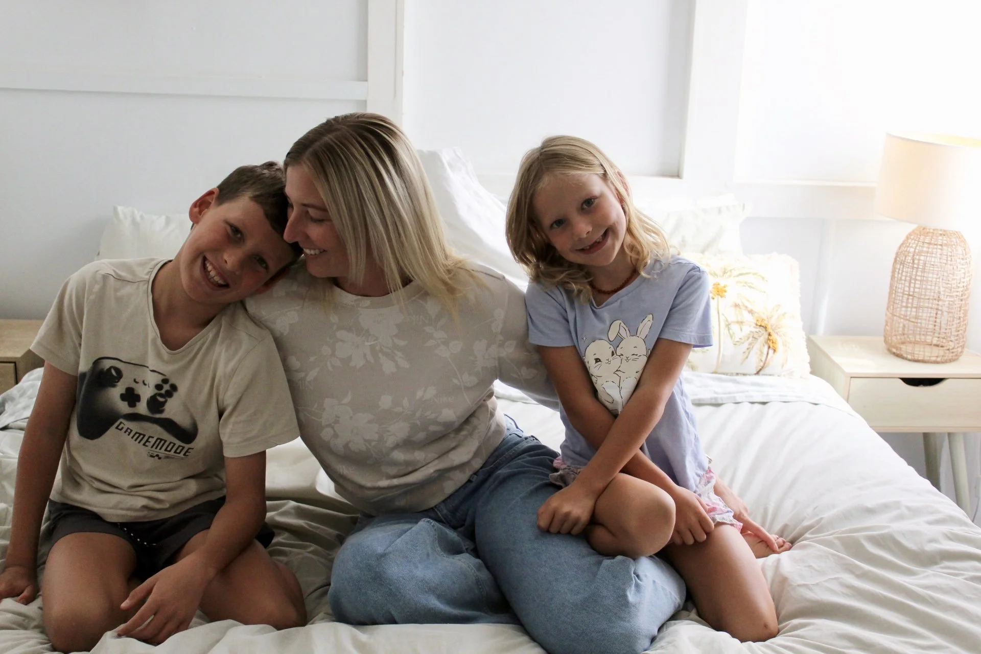 A woman with blonde hair sitting on a bed with a boy and a girl, all smiling and leaning their heads against each other. The boy is wearing a t-shirt with a game controller graphic, and the girl is wearing a light-colored shirt with bunny illustrations. The room has white walls and bedding, with a nightstand and lamp beside the bed.