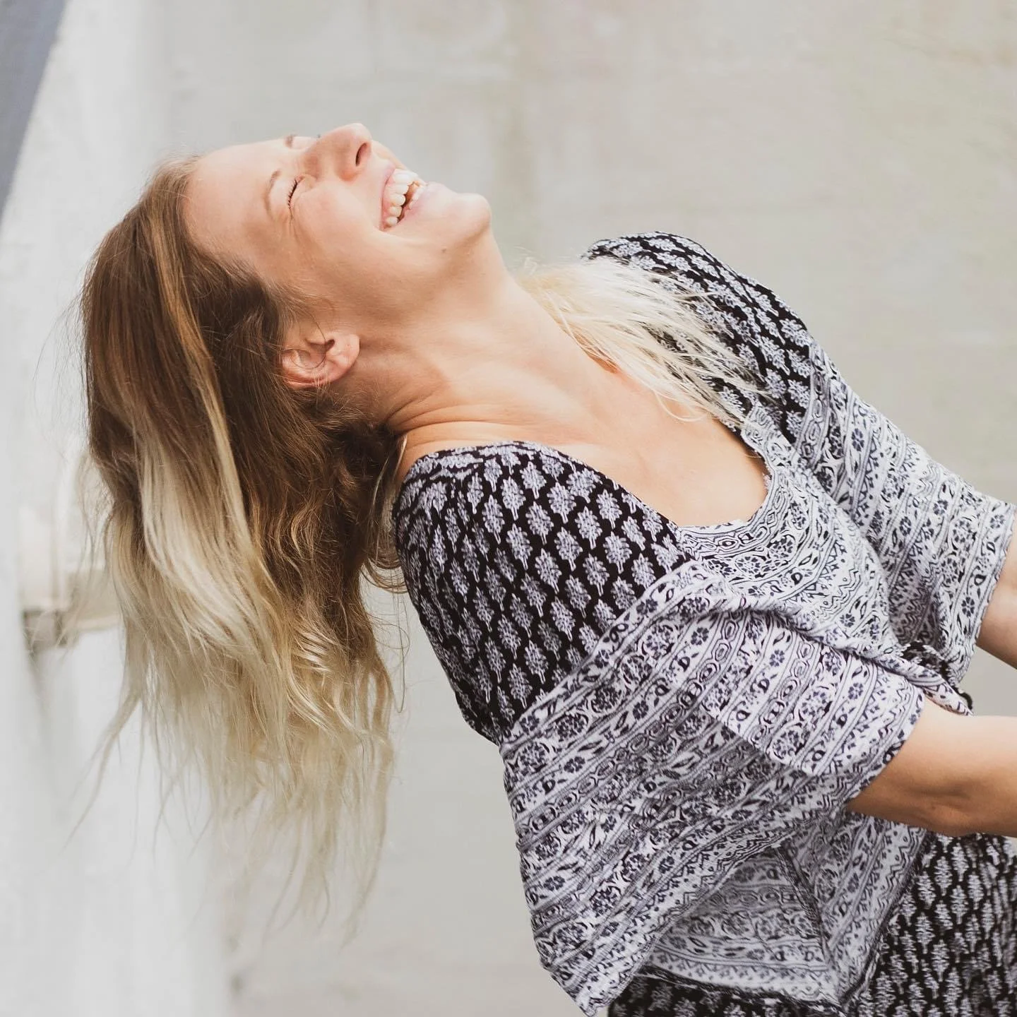 A woman with long blonde hair smiling and leaning back, wearing a patterned blue and white dress.