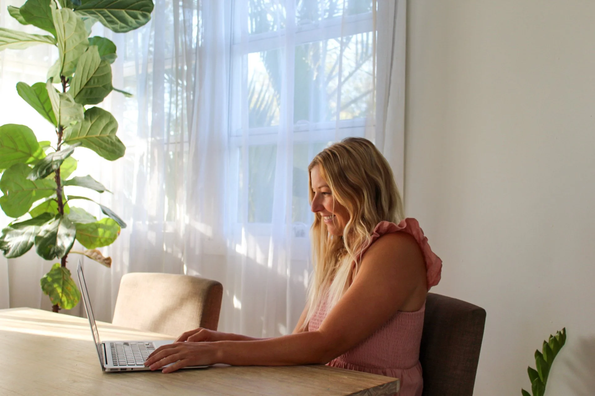 A woman with long blonde hair, smiling and working on a laptop at a table in a bright room with white curtains and a large green plant to her left.