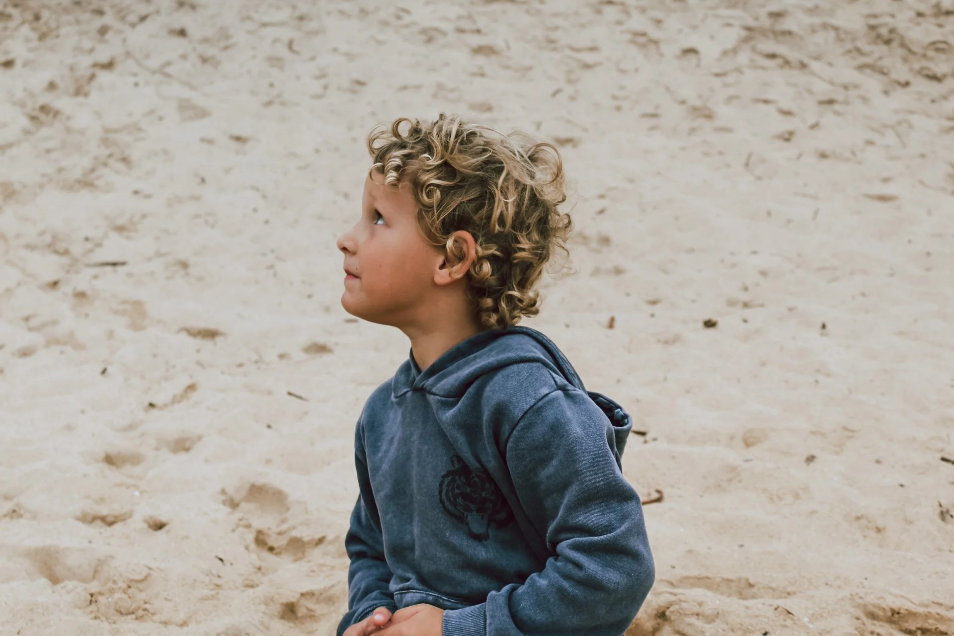 A young boy with curly blond hair sitting on a sandy beach, looking to his left.