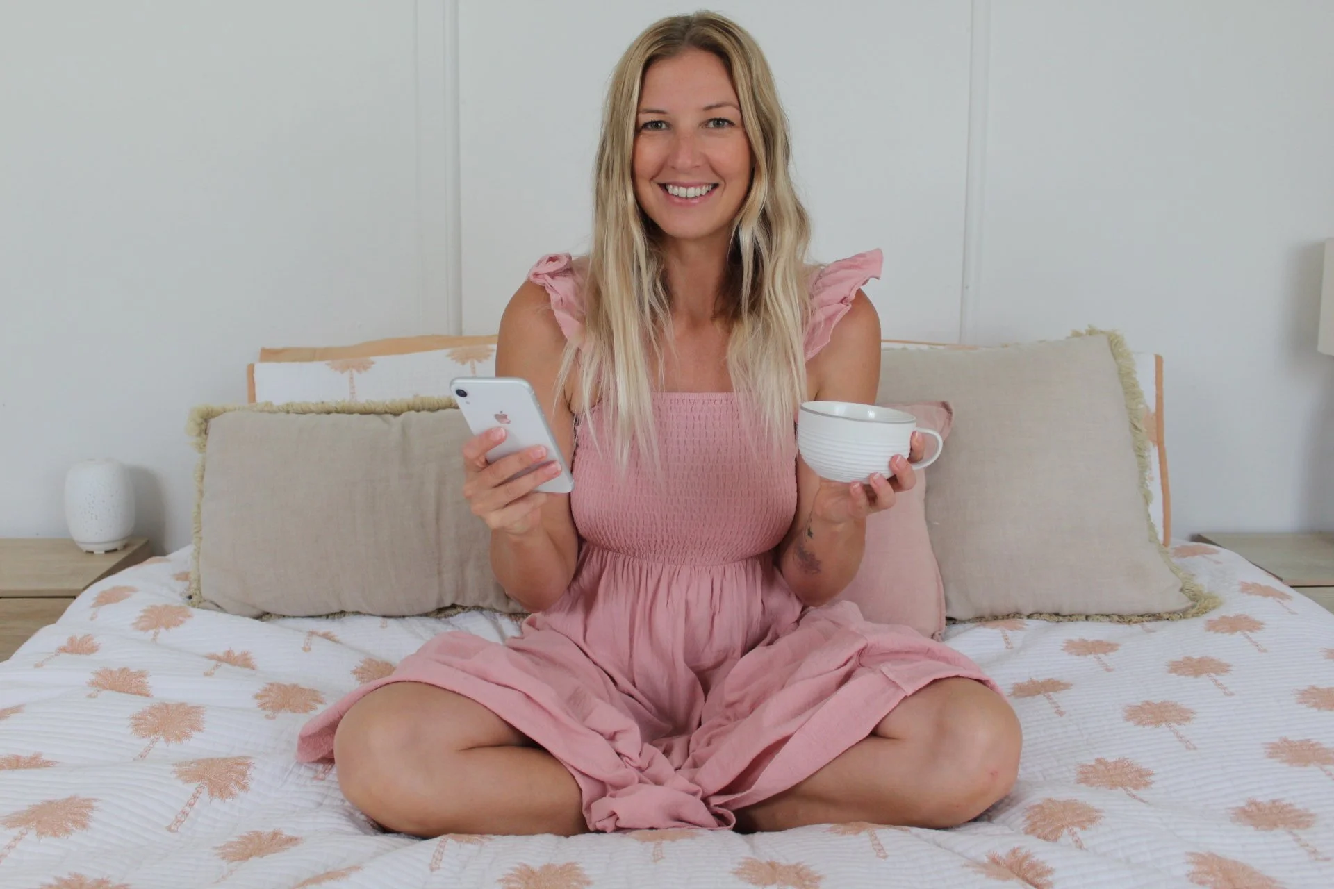 A woman with long blonde hair, wearing a pink dress, is sitting cross-legged on a bed in a bedroom. She is smiling and holding a white smartphone in her right hand and a white mug in her left hand. The bed has beige pillows with fringed edges and a white bedspread with a pink floral pattern. There is a small white lamp on a bedside table on the left side of the image and a white wall in the background.