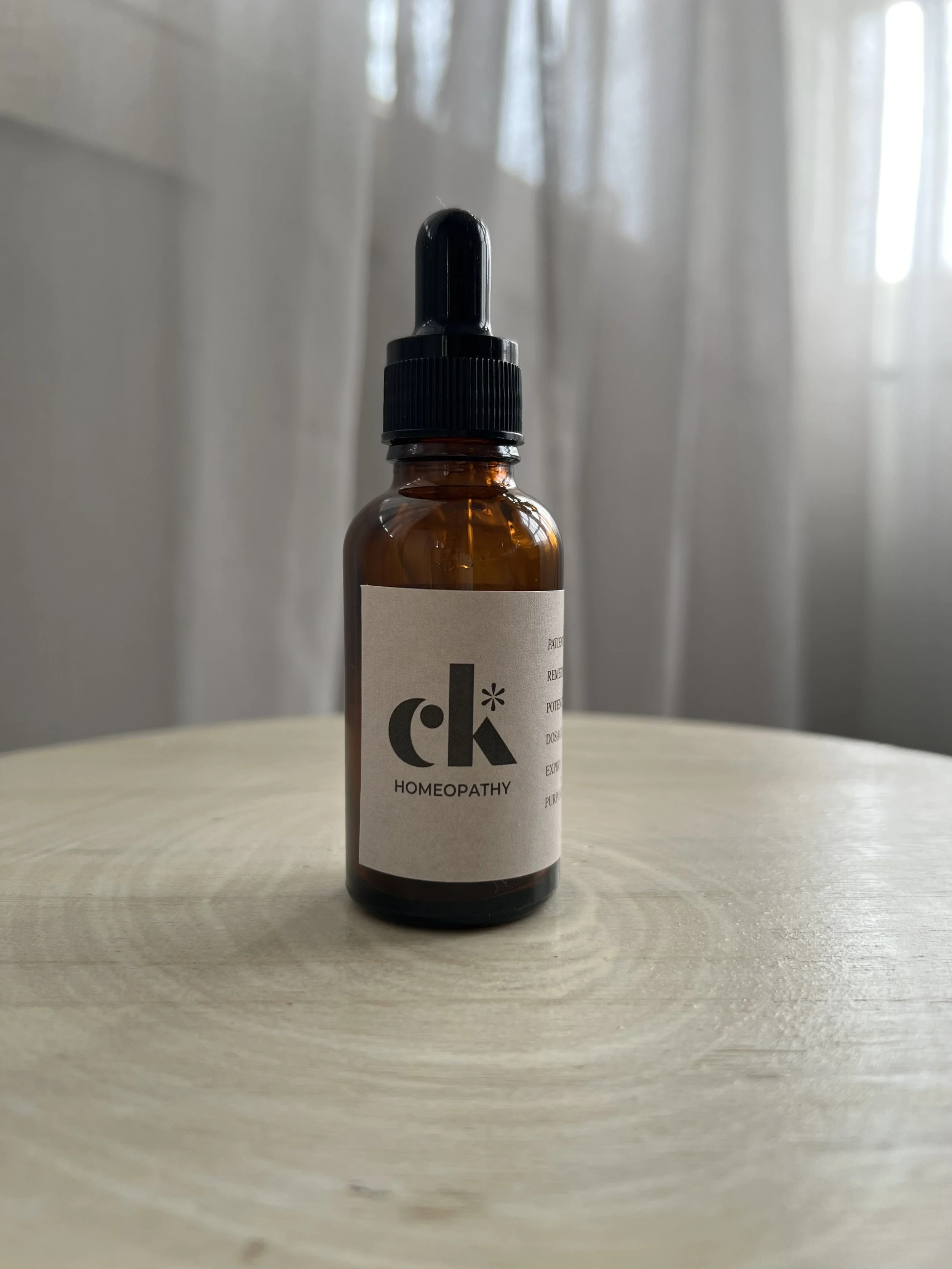 A brown glass bottle with a black dropper cap labeled 'ck* HOMEOPATHY' placed on a light wooden surface with a blurred background and white curtains.