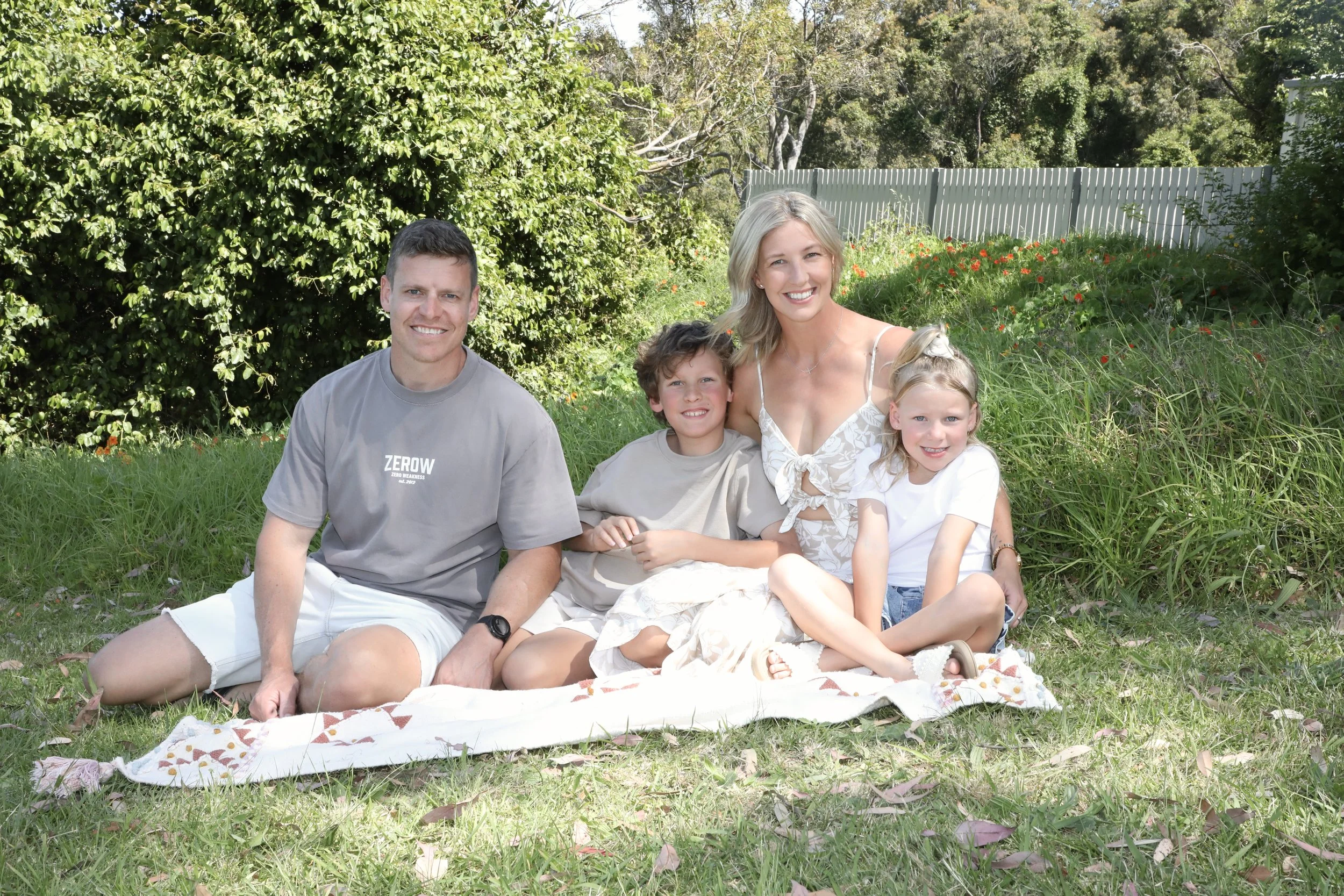 A family of five sitting on a blanket outdoors in a sunny backyard. The family includes a man, a woman, two girls, and a boy, all smiling and looking at the camera. The background features green bushes, trees, and a white fence.