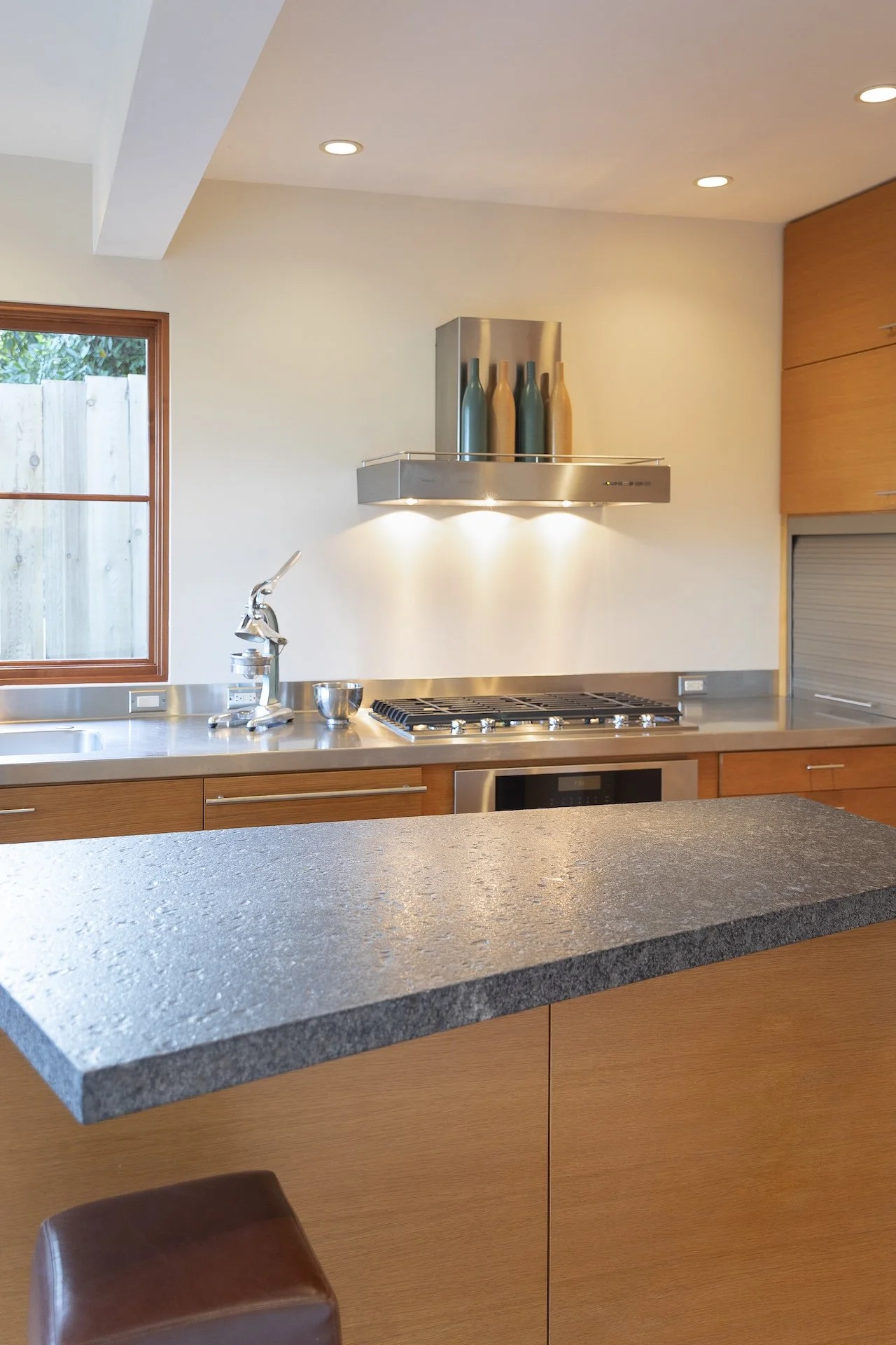 A modern kitchen with a granite countertop, wooden cabinets, a stainless steel range hood with decorative bottles, a faucet, and a window showing an outdoor fence.