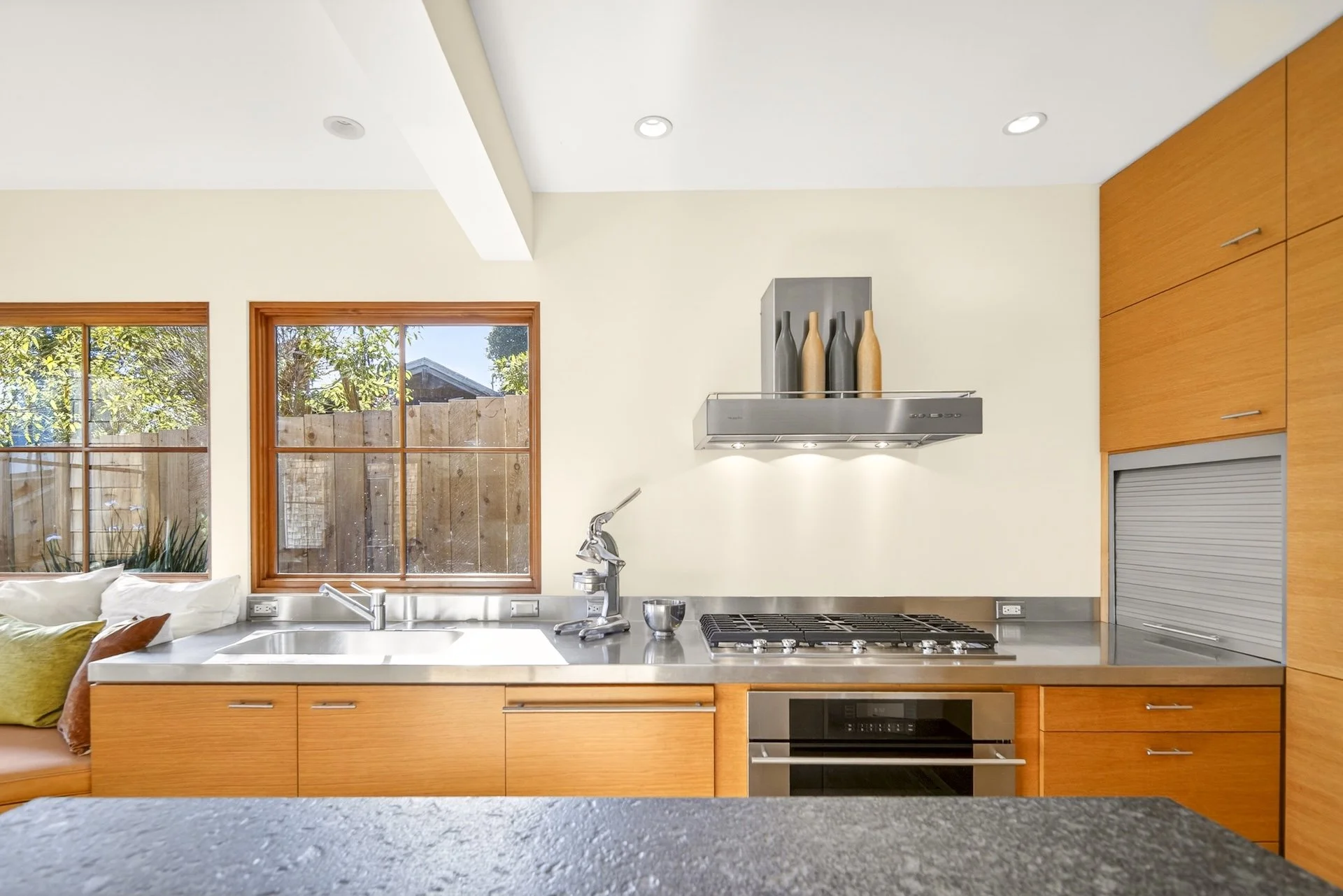 Modern kitchen with wooden cabinets, stainless steel appliances, and large windows showing trees outside.