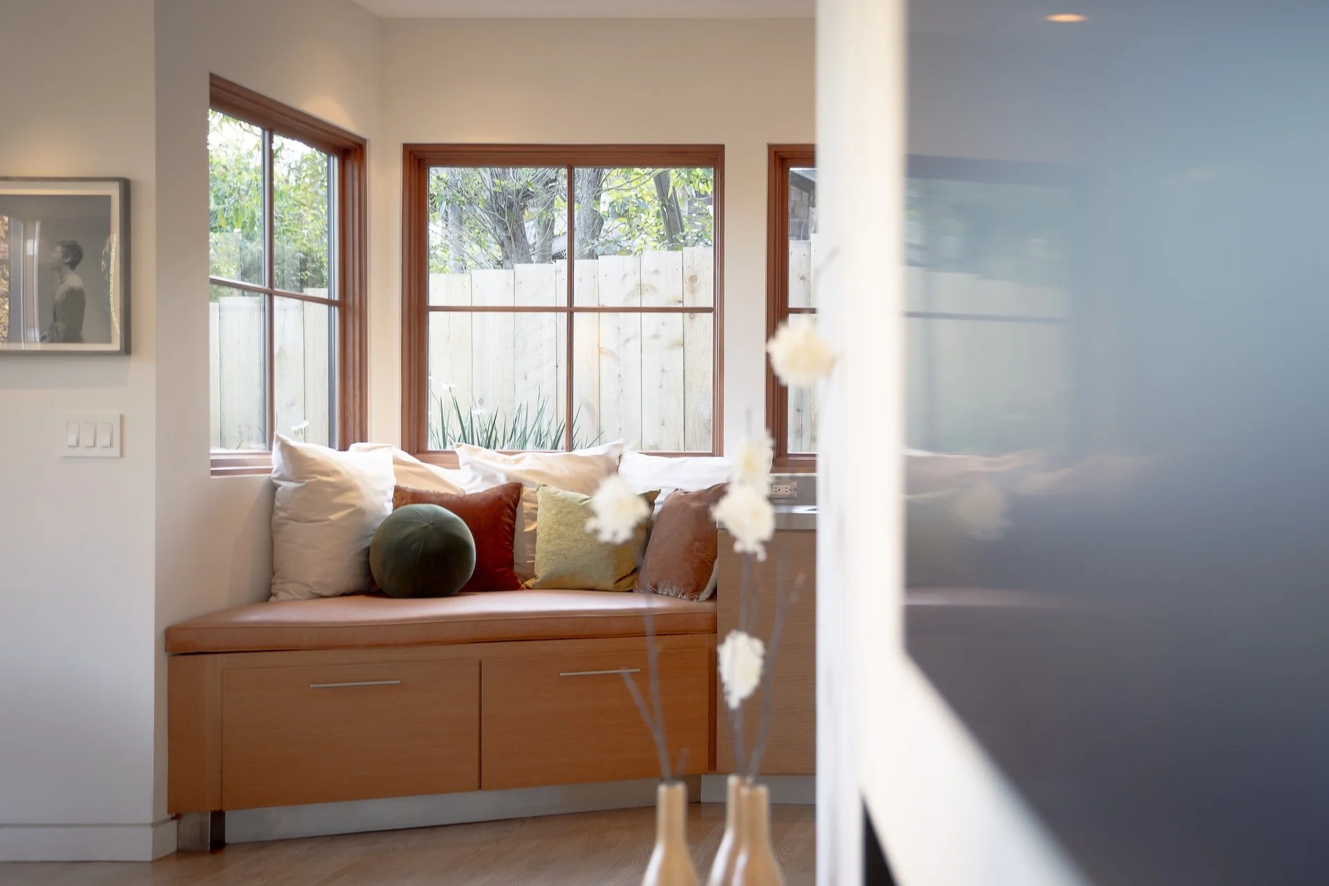 Interior of a cozy living space with a built-in wooden bench seat topped with white, beige, green, and rust-colored pillows. Large windows with wooden frames reveal a backyard with trees and a wooden fence, allowing natural light to fill the room. A 