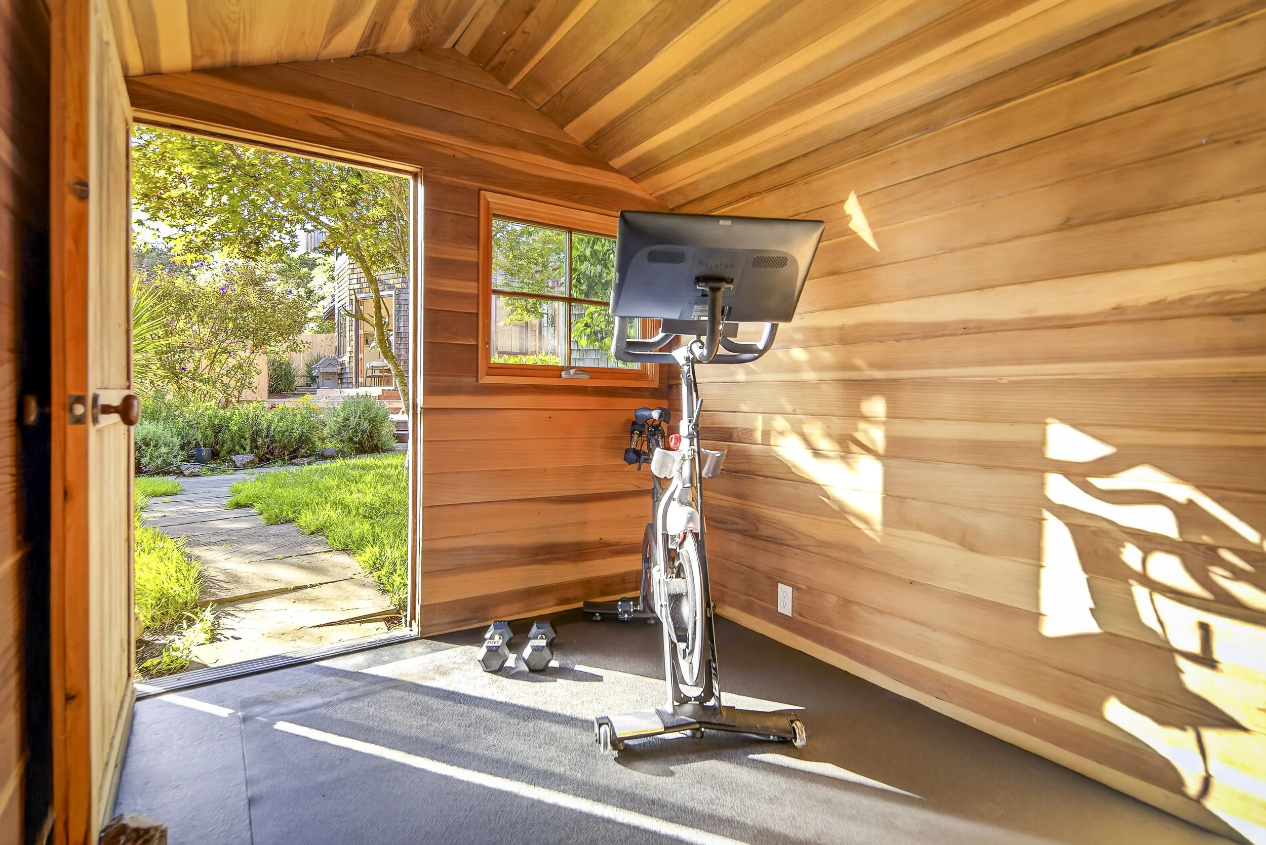 A wooden interior room with a stationary exercise bike and a small set of dumbbells on the floor, with large windows showing a lush green garden outside.