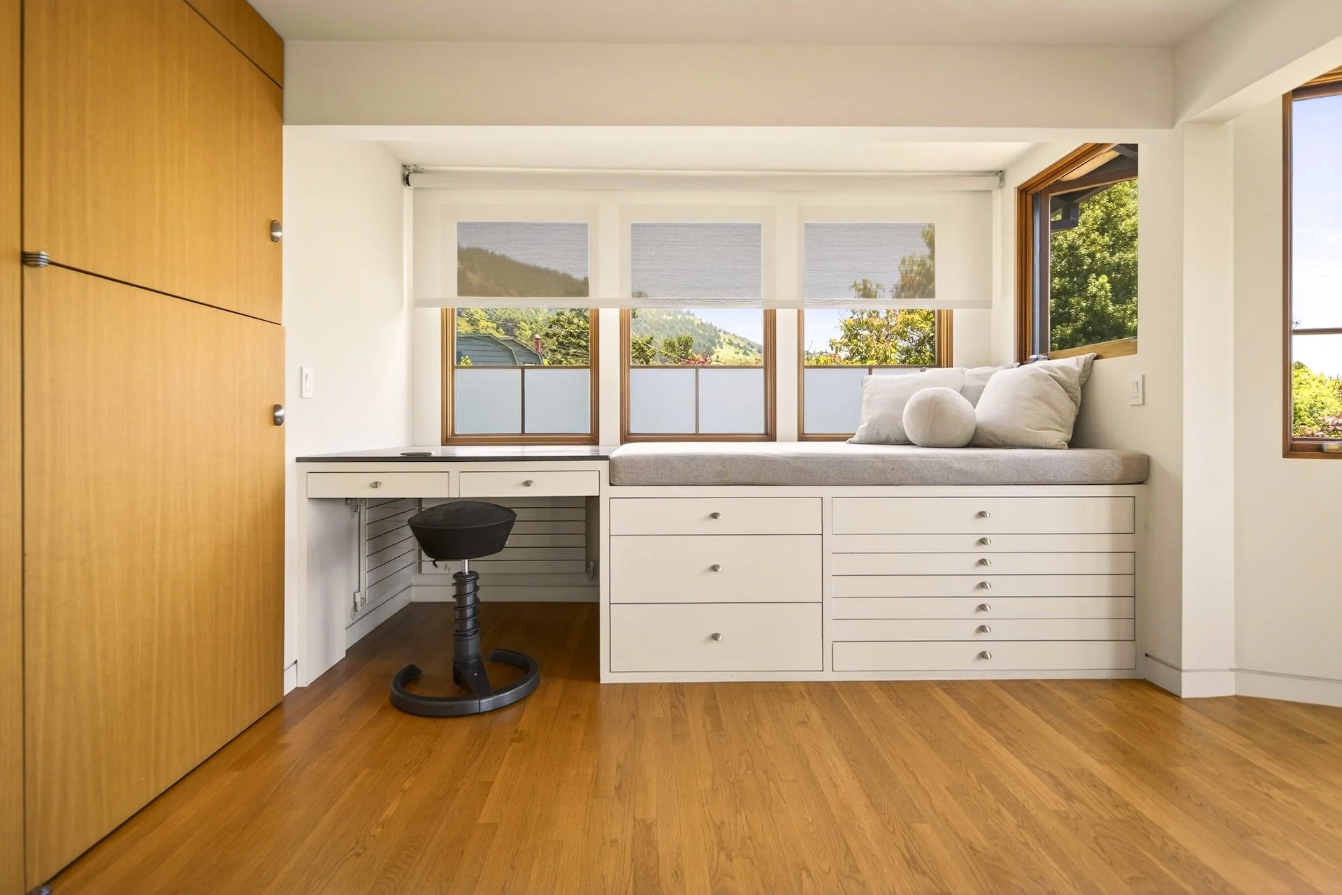 A bright, modern home office corner with a white built-in desk and drawers, a black swivel chair, and large windows with views of greenery and trees outside.