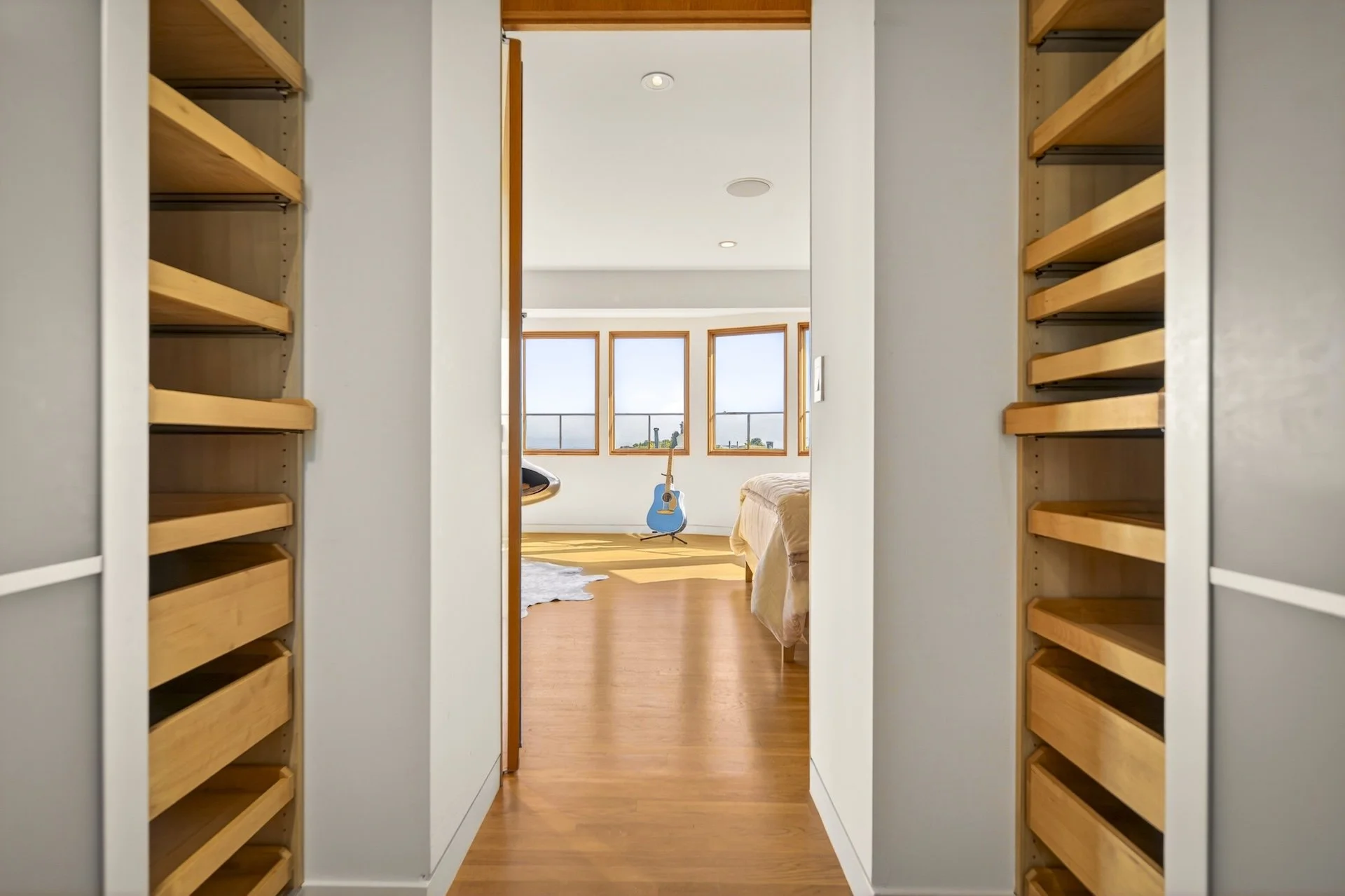View through a walk-in closet opening into a bedroom with large windows, a blue guitar, a bed, a white rug, and hardwood flooring.