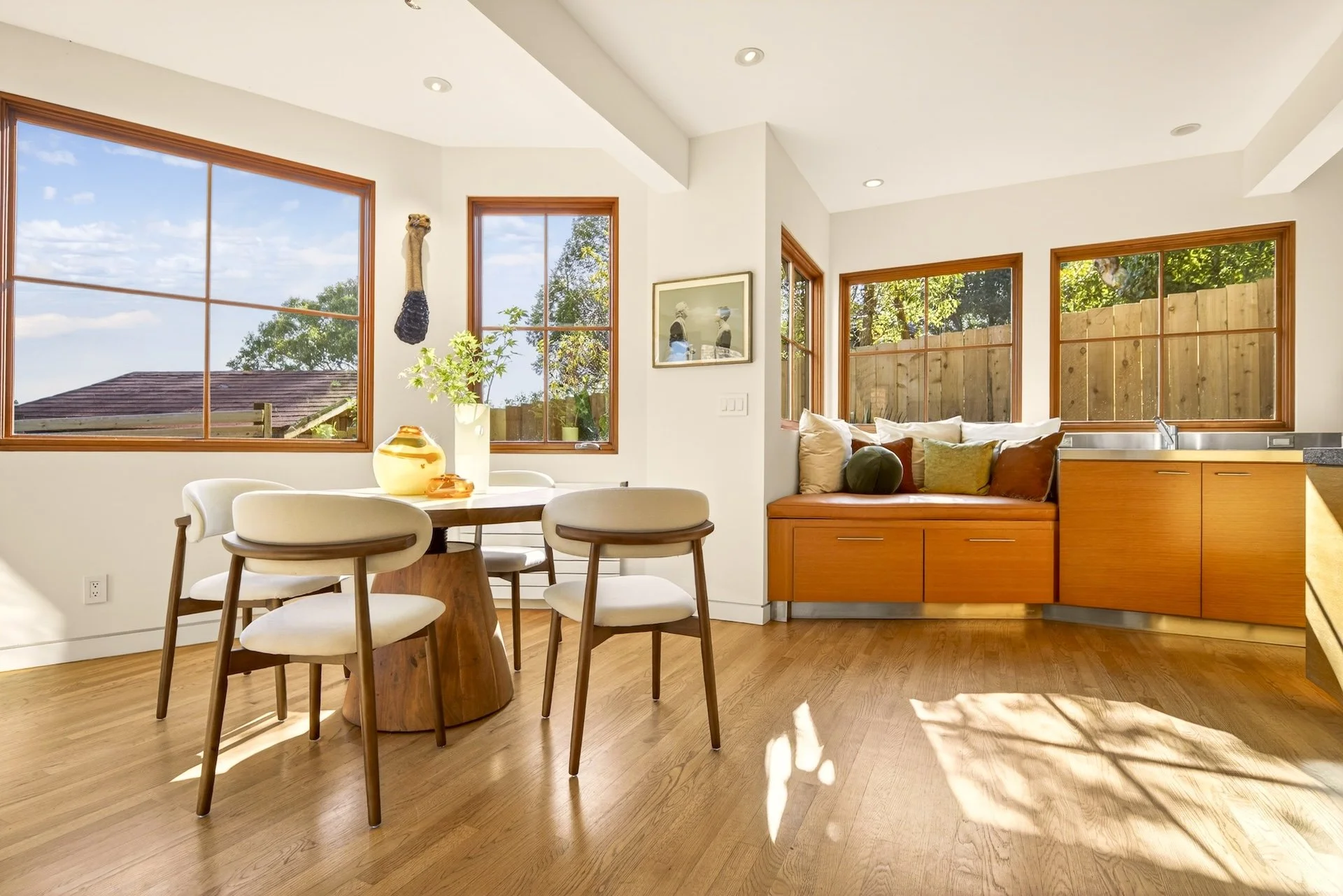 Bright and airy dining and kitchen area with large windows, a round wooden dining table with white chairs, and a window seat with pillows under natural sunlight.