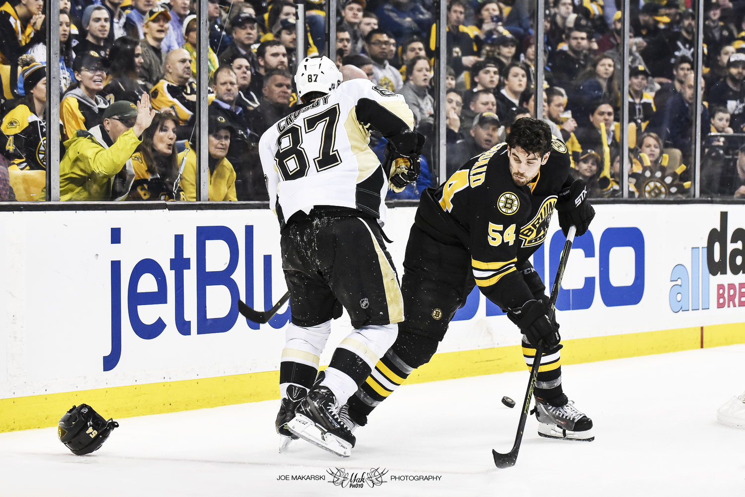 Two hockey players, one from the Boston Bruins wearing black and yellow and the other from the Pittsburgh Penguins in white and black, compete for the puck along the rink's sideboards during a game. The crowd watches from behind the glass.