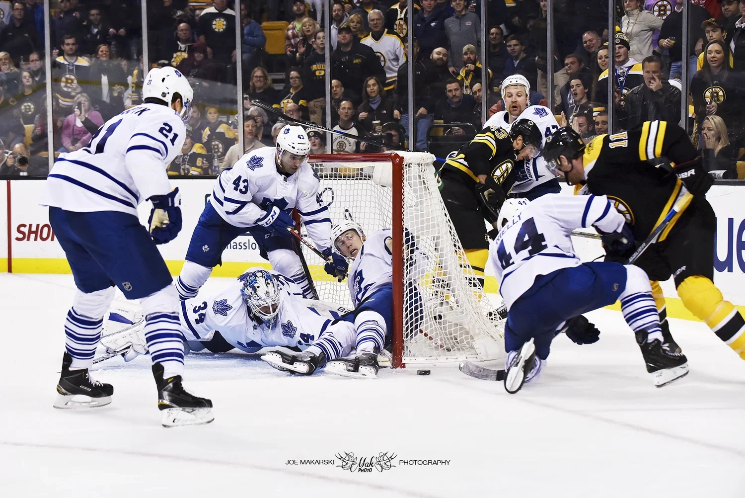 Hockey players from the Toronto Maple Leafs and Boston Bruins in a tense moment near the goal during a game, with the puck loose on the ice and spectators watching from the stands.