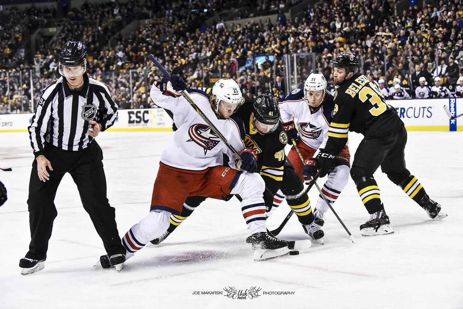 Hockey players from two teams competing for the puck during a game, with an official observing nearby in an arena filled with spectators.