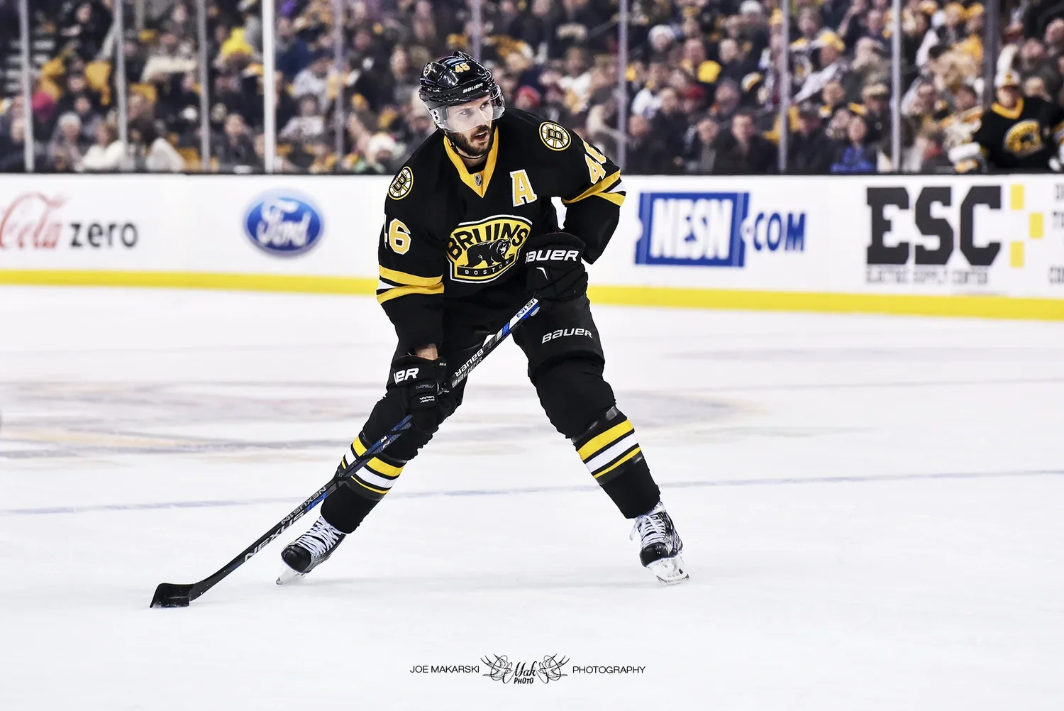 A hockey player in black and yellow Boston Bruins jersey with the number 46, wearing black skates and holding a hockey stick, on the ice during a game.