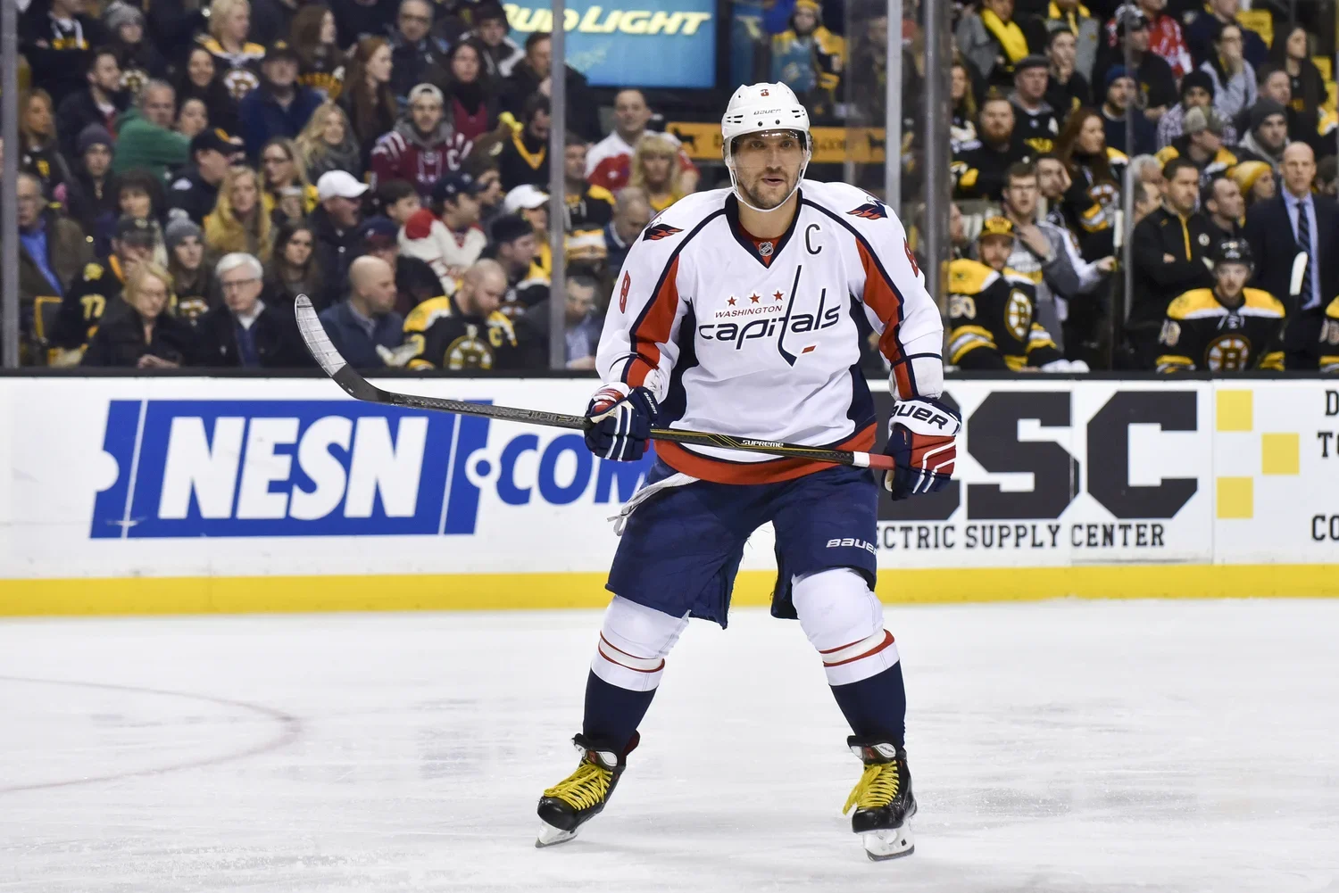 An ice hockey player from the Washington Capitals in uniform standing on the ice with a hockey stick, during a game with fans in the background.