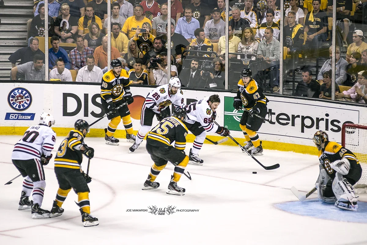 Hockey players from opposing teams competing near the goal during a game, with spectators watching from the stands.