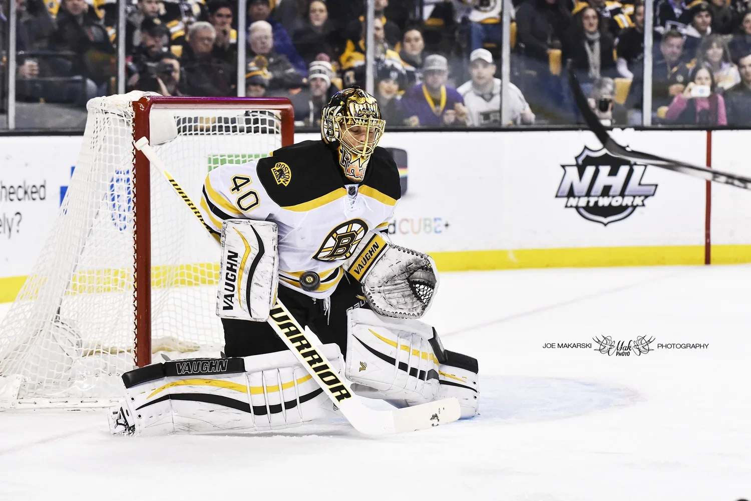 Ice hockey goaltender in black and white uniform with yellow accents kneeling in front of the net during a game, with a crowd watching behind him.