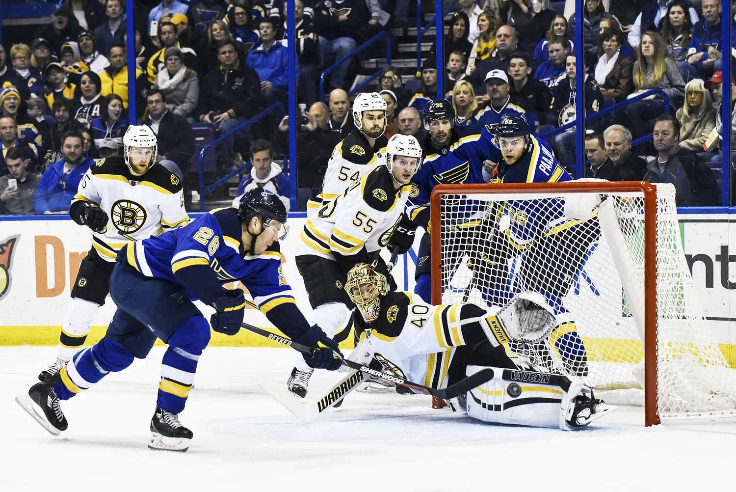 Ice hockey game with players from opposing teams, one team in blue jerseys and the other in white jerseys, near the goal. A player in blue attempts to score, while the goalie in white and yellow pads blocks the puck. Fans are in the background watchi